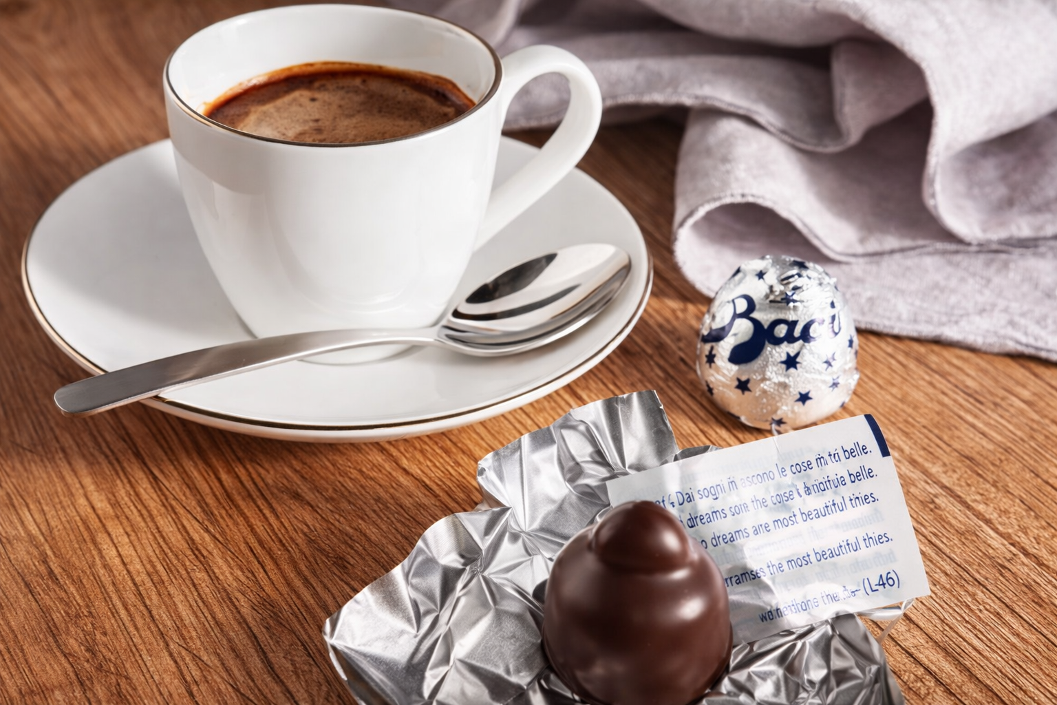 A white cup of coffee on a saucer with a teaspoon, a wrapped chocolate candy, a foil wrapper, and a small foil-wrapped chocolate egg on a wooden surface with a gray cloth in the background.