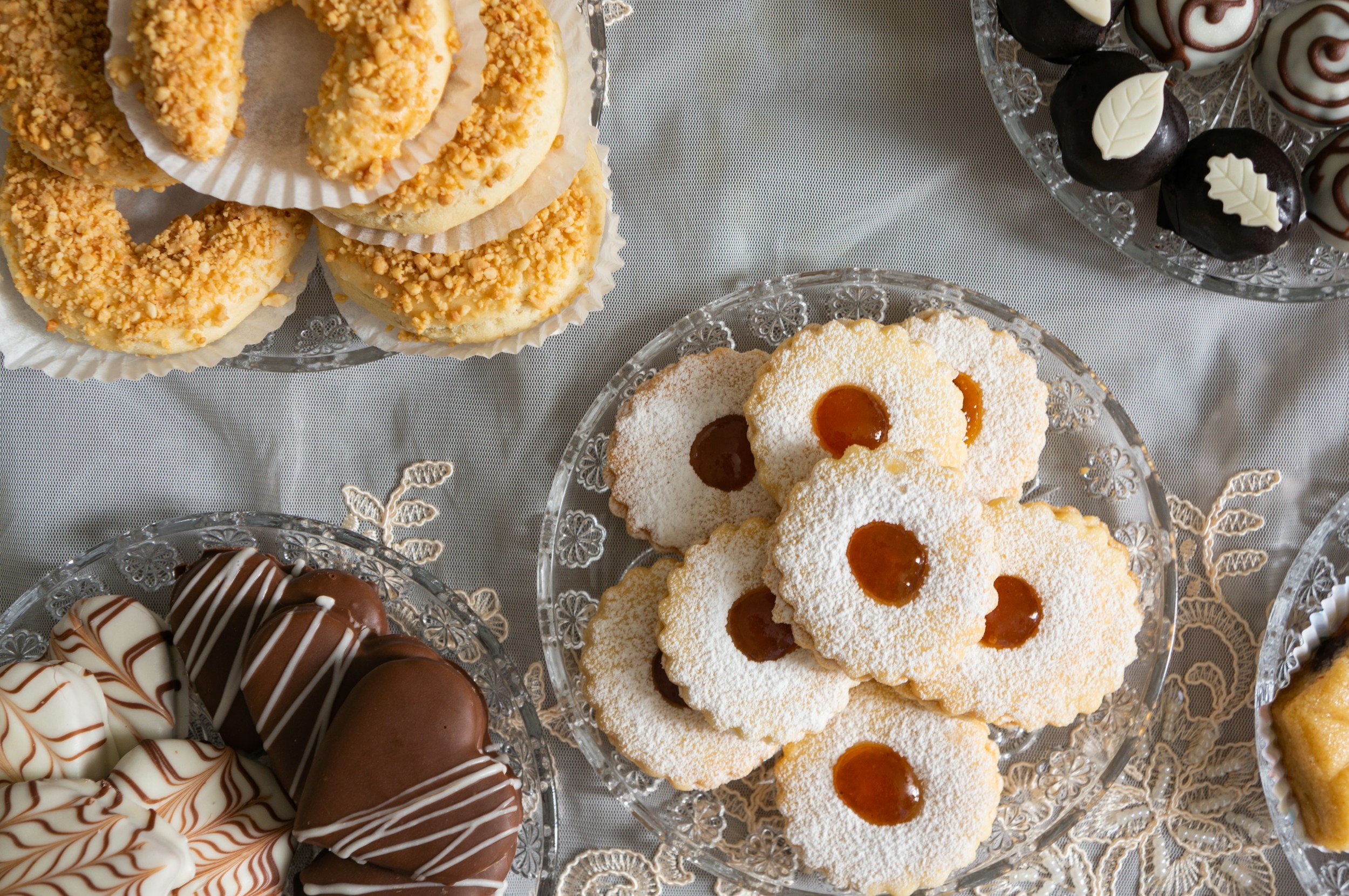 Various decorated cookies and donuts on crystal glass plates, arranged on a white tablecloth.