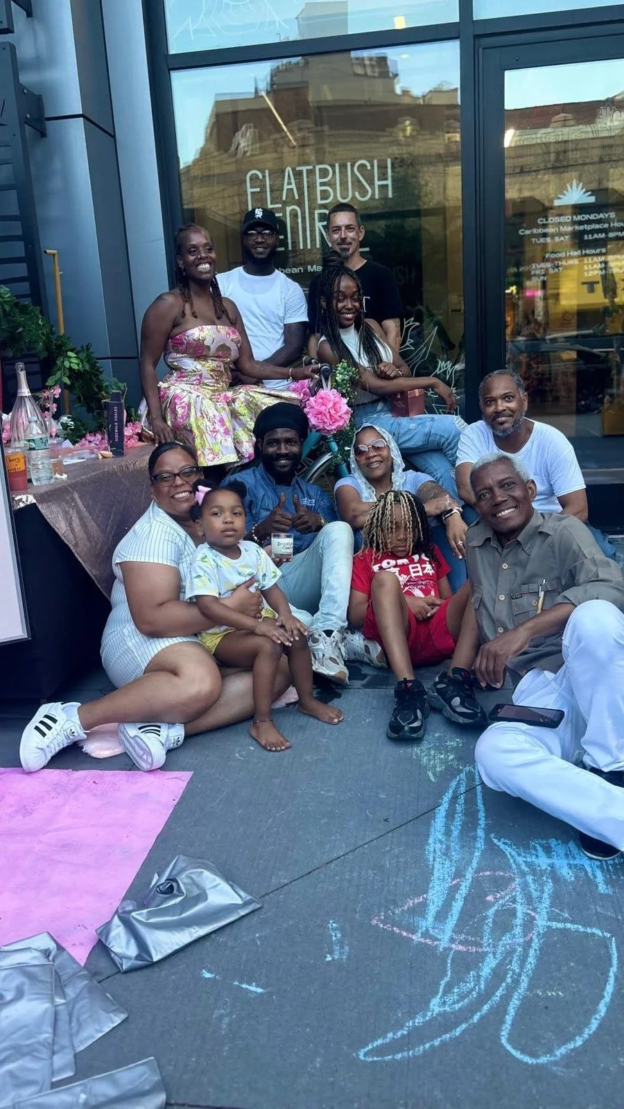 Group of people gathered outside a building with the sign 'Flatbush' and floral decorations. Some are sitting on the ground, others standing or sitting on a bicycle, smiling and enjoying a social moment.