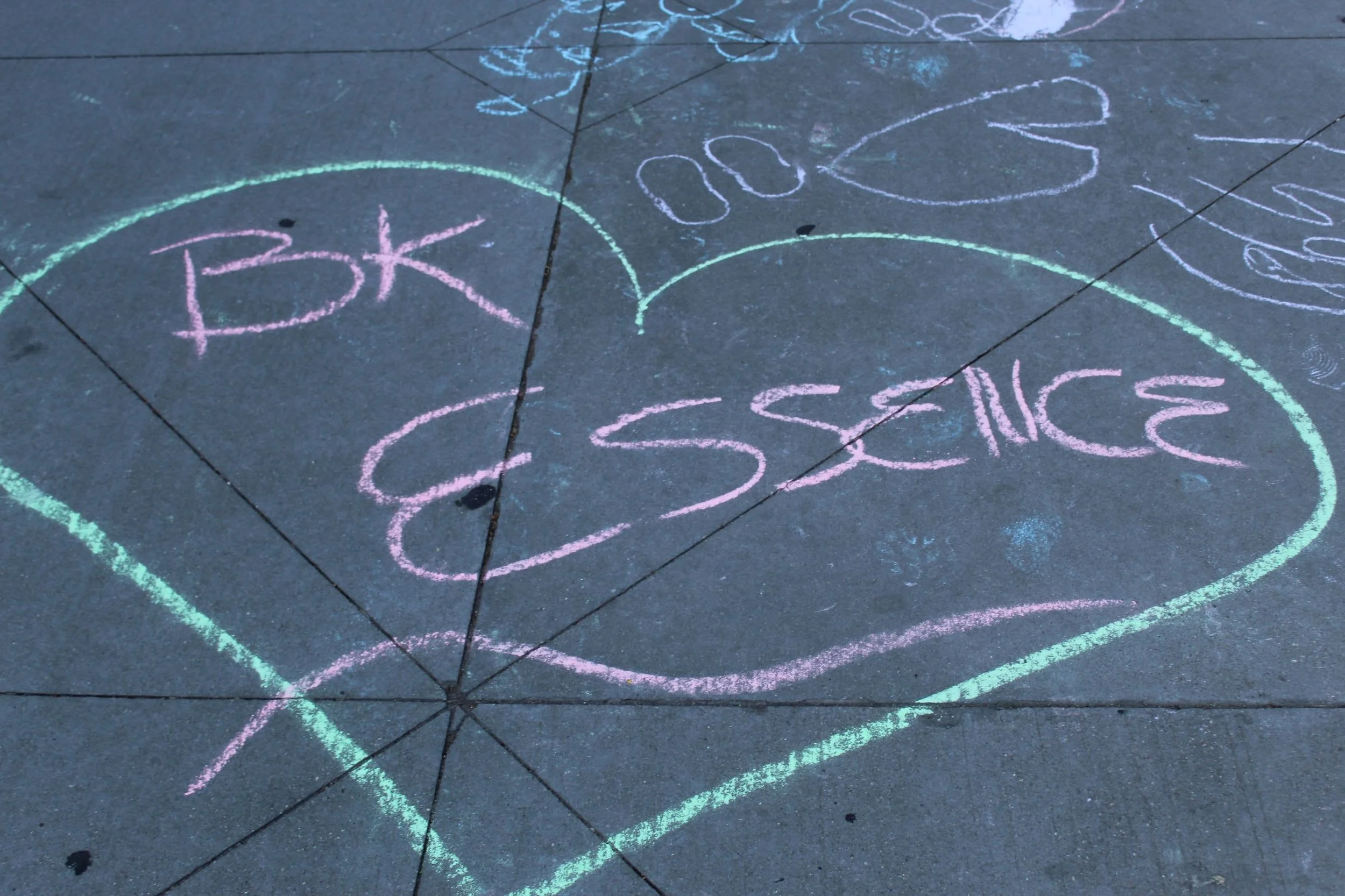 Colorful chalk drawing on concrete pavement with words "BE KIND" enclosed in a heart shape, along with other chalk doodles and writing.