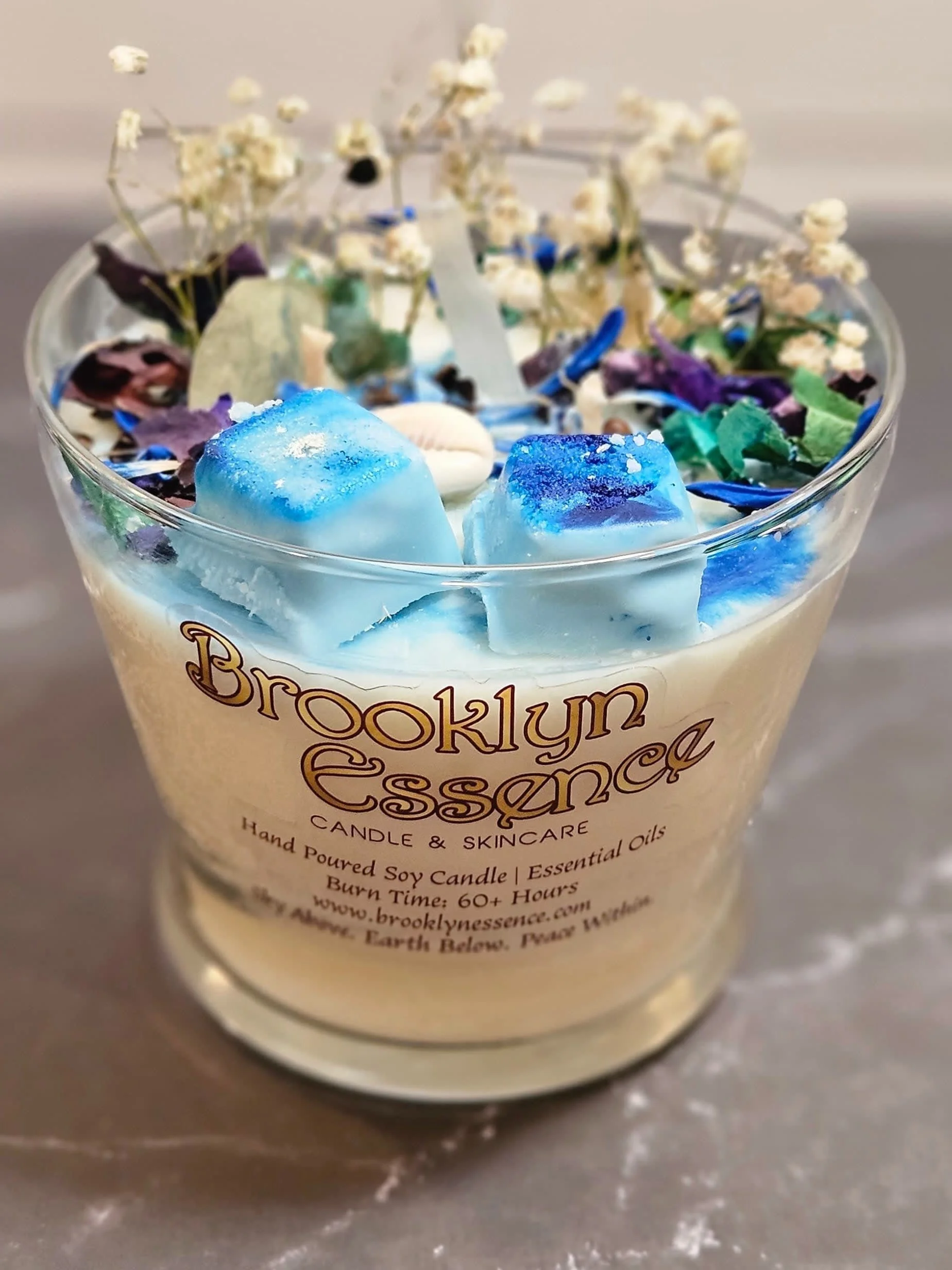 Close-up of a Brooklyn Essence candle with decorative blue and white wax melts, dried flowers, and colorful dried leaves inside a glass jar.