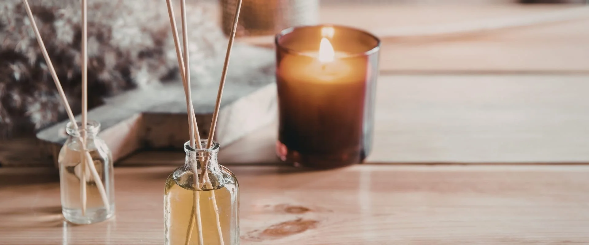 Close-up of small glass bottles with reed diffusers and a lit candle in a glass holder on a wooden surface.