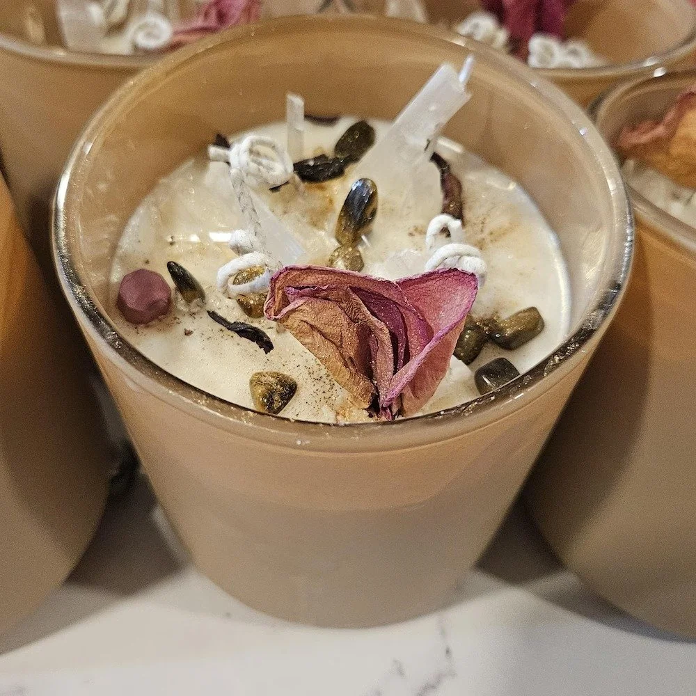 A beige candle in a glass container decorated with dried rose petals, green seeds, and small white and pink objects.