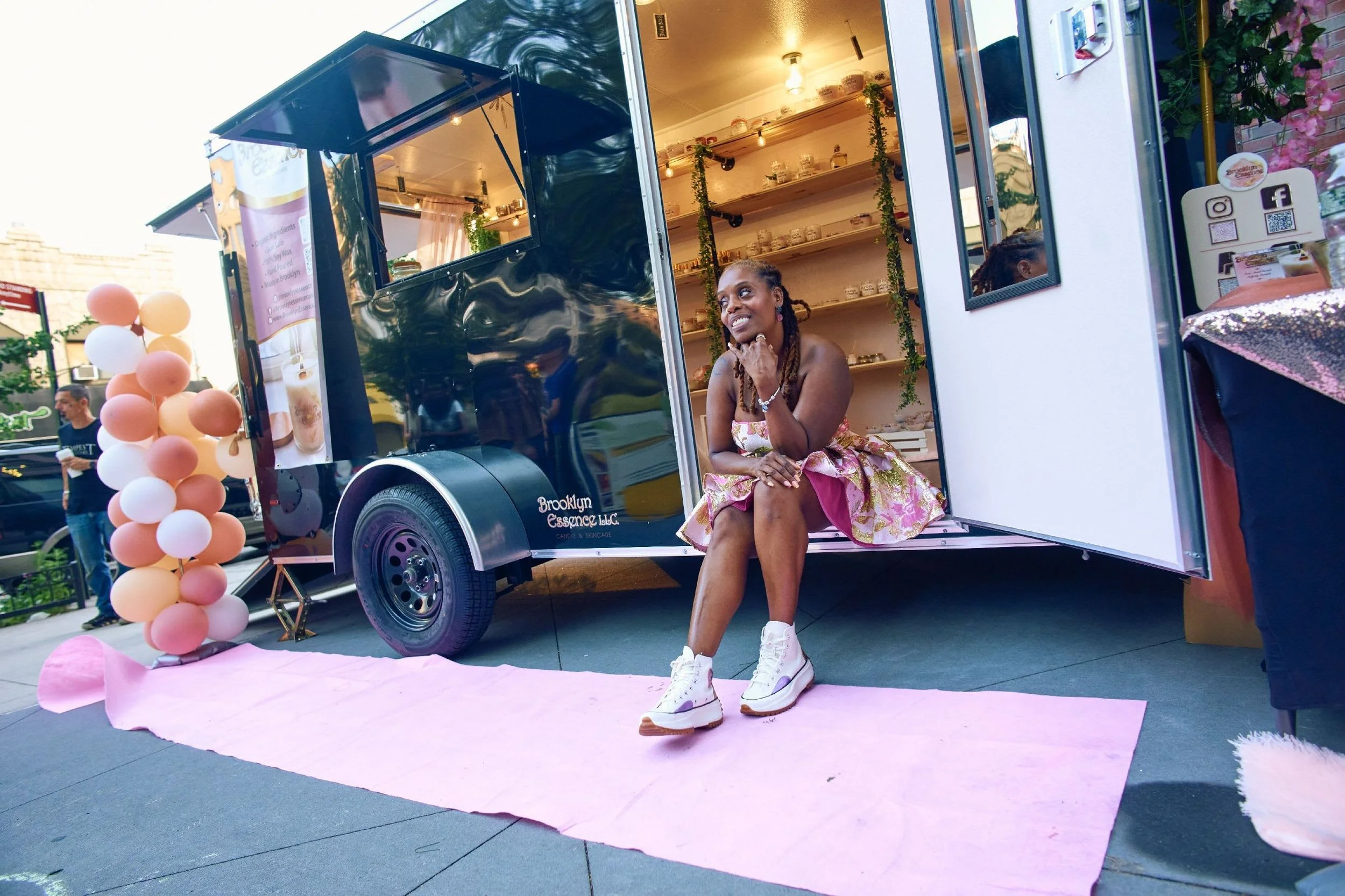 A woman sitting on the step of a black food truck, smiling and looking to the side. She is wearing a floral dress with white sneakers. The food truck has an open window and wooden shelves inside. Pink and peach balloonsDecorate the scene, and a pink cloth is spread on the ground in front of her.