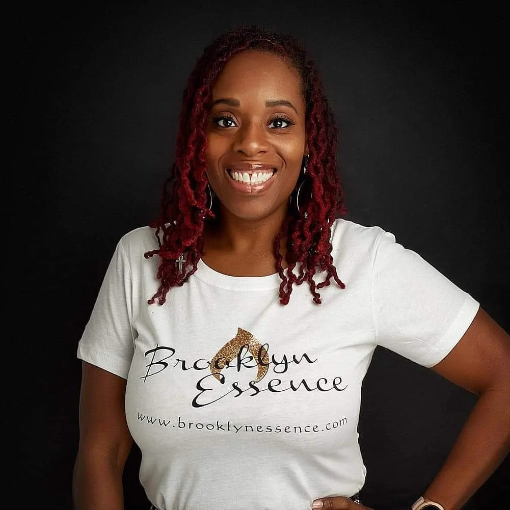 A woman with red dreadlocks smiling, wearing a white t-shirt that says 'Brooklyn Essence' and features a logo with a gold-colored circle, standing against a black background.