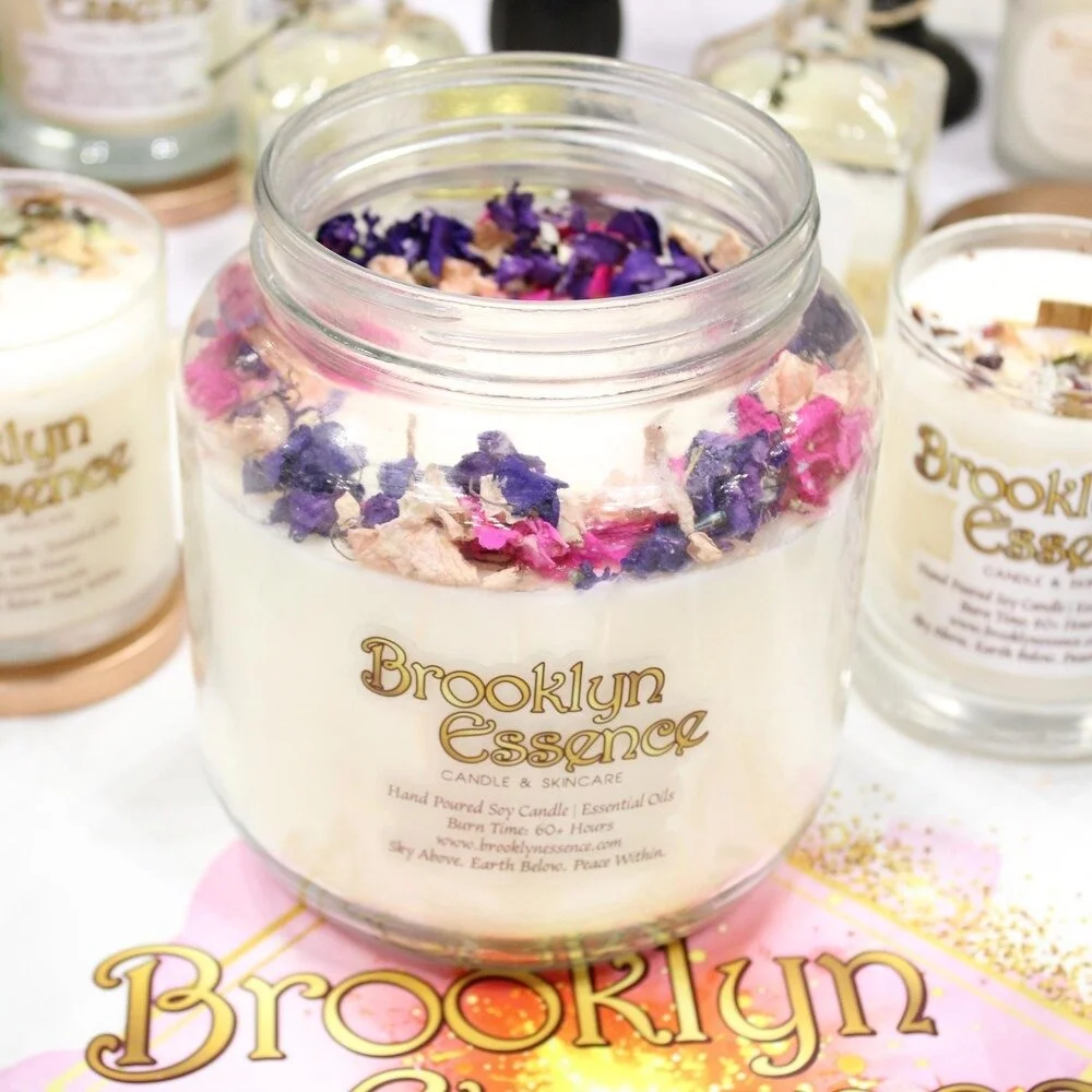 A large glass jar candle labeled 'Brooklyn Essence' with dried purple, pink, and beige flower petals on top. Several other candles are visible in the background.