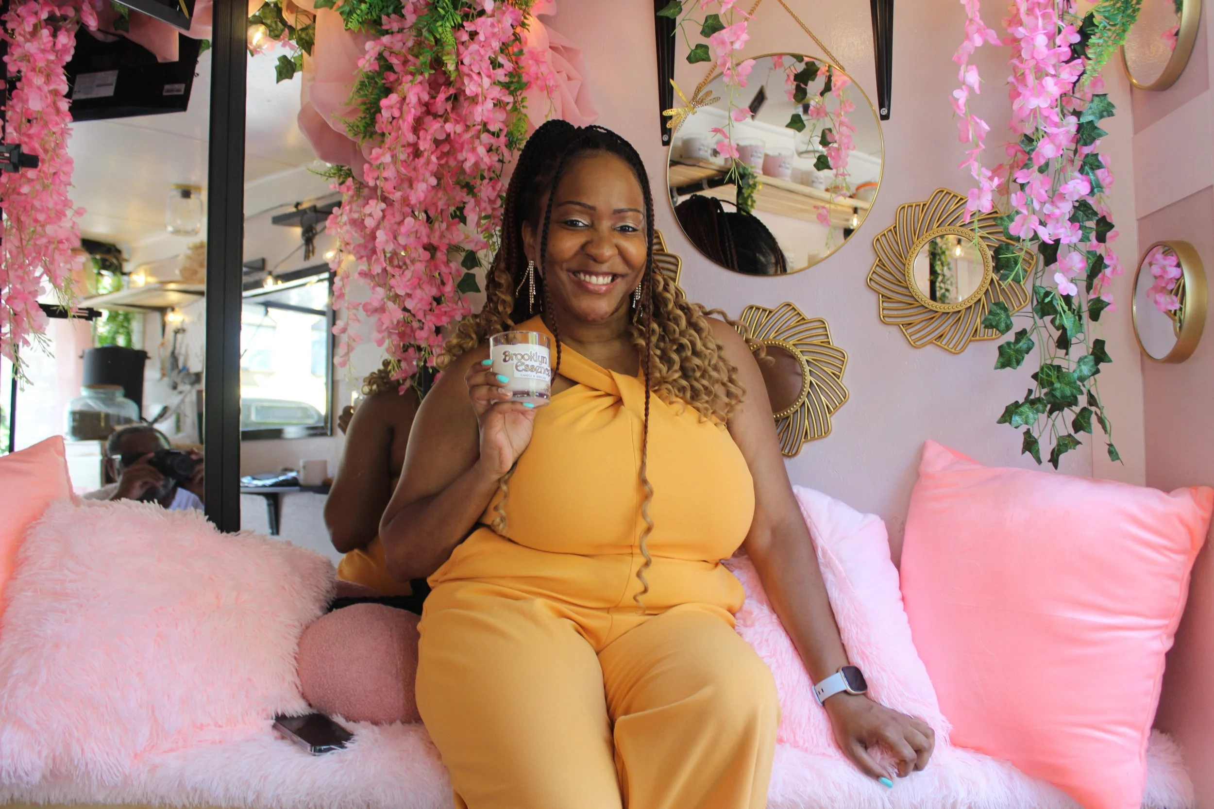 Smiling woman with long braided hair, wearing a yellow jumpsuit, sitting on a pink fluffy couch with pink pillows, holding a cup that says 'Brooklyn ESSA' inside a decorated boutique or cafe with pink flowers and gold mirror accents on the wall in th