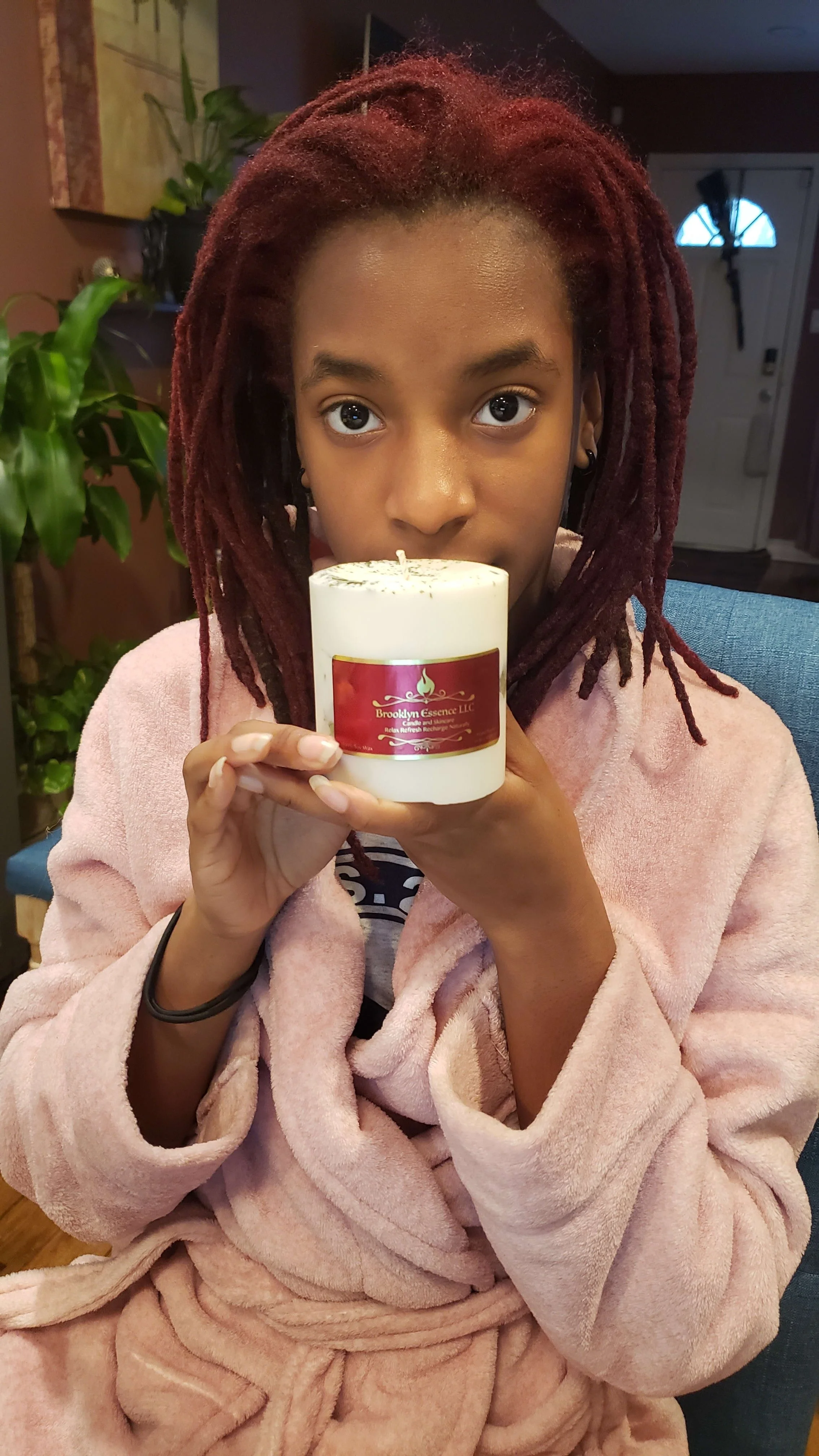 A young girl with red dreadlocks wearing a pink bathrobe holding a white candle with a red label, indoors with a green plant and a door in the background.