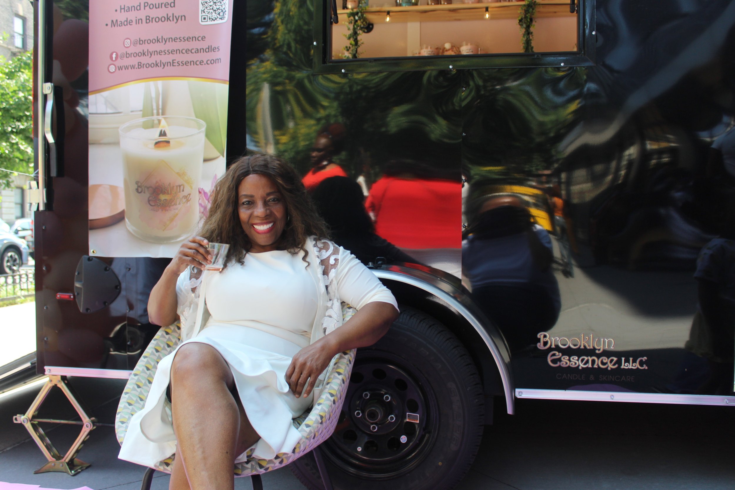 A woman in a white dress sitting on a chair outside, smiling and holding a small cup, next to a black food truck with a sign for Brooklyn Essence LLC. The sign features an image of a candle and the company’s social media handles.