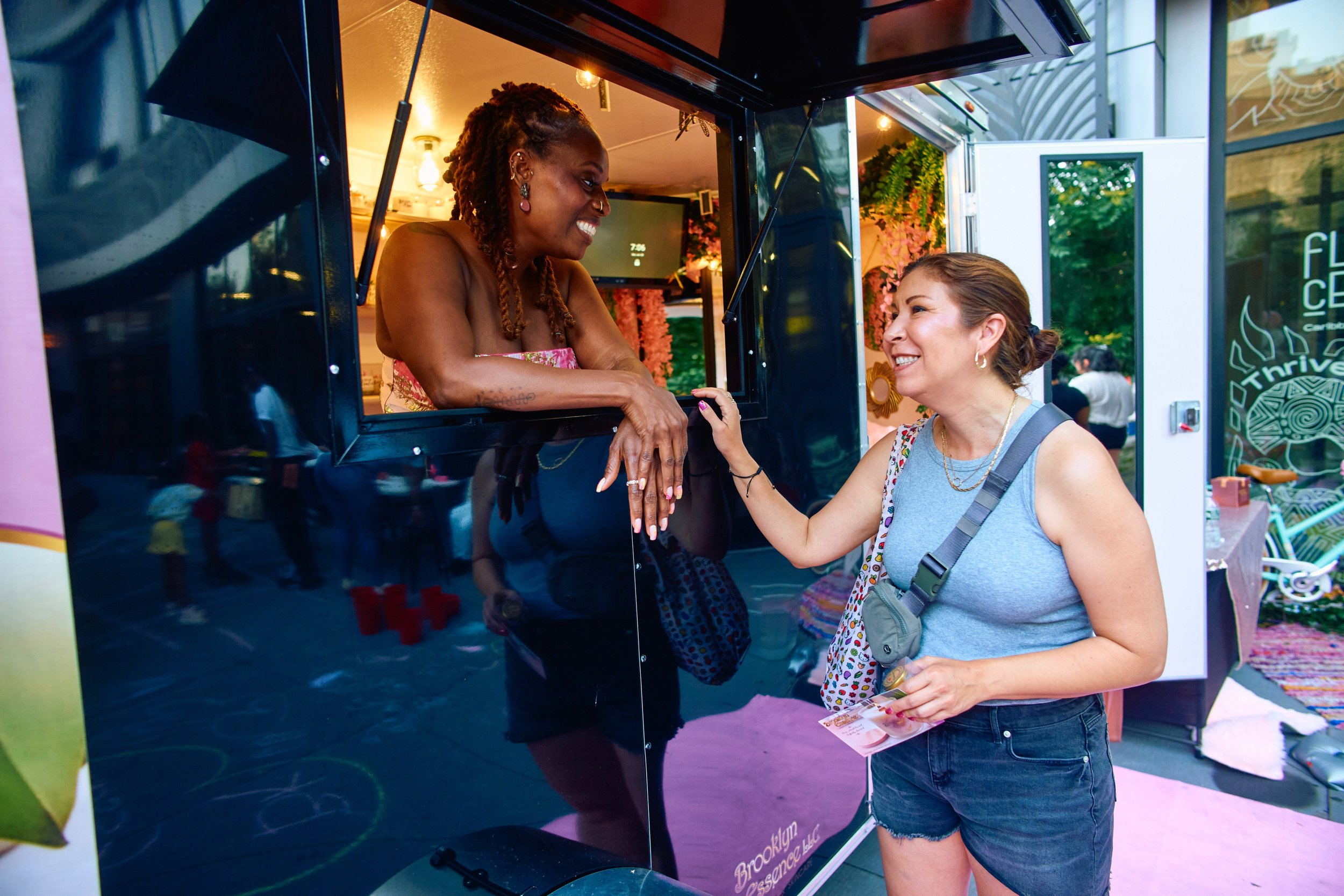 Two women interacting at a food truck or kiosk, smiling and engaging in conversation. One woman, with braided hair, leans out of the truck window and is wearing a strapless top. The other woman, with short brown hair, is holding money and a travel ca