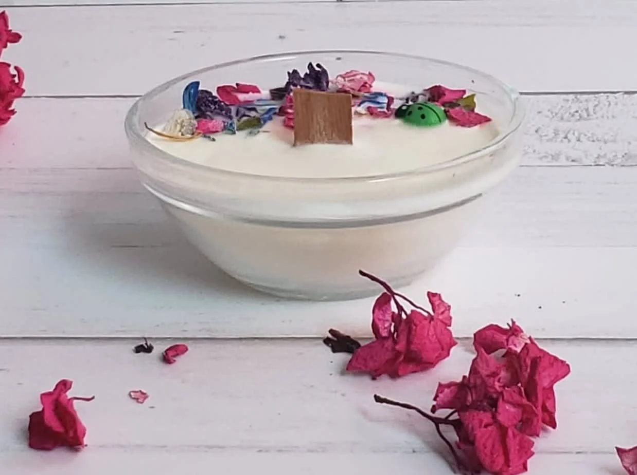 A glass bowl filled with creamy white pudding topped with colorful sprinkles, flower petals, and small decorative items on a white wooden surface with some pink dried flowers scattered around.