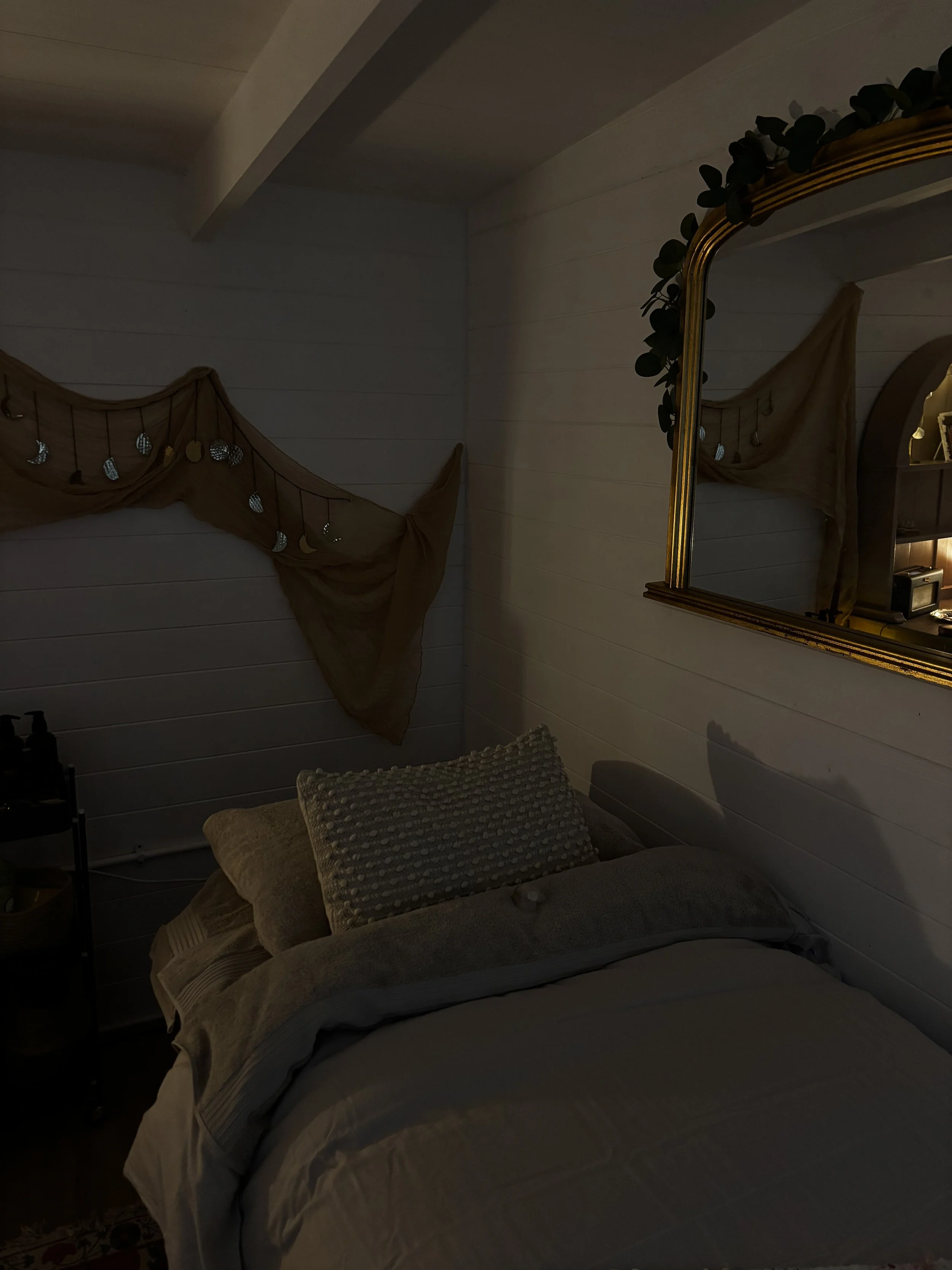 Cozy bedroom with a bed decorated with pillows and a blanket, a mirror on the wall with green foliage on top, fairy lights and moon decoration on a brown draped fabric, and a small side table with bottles.