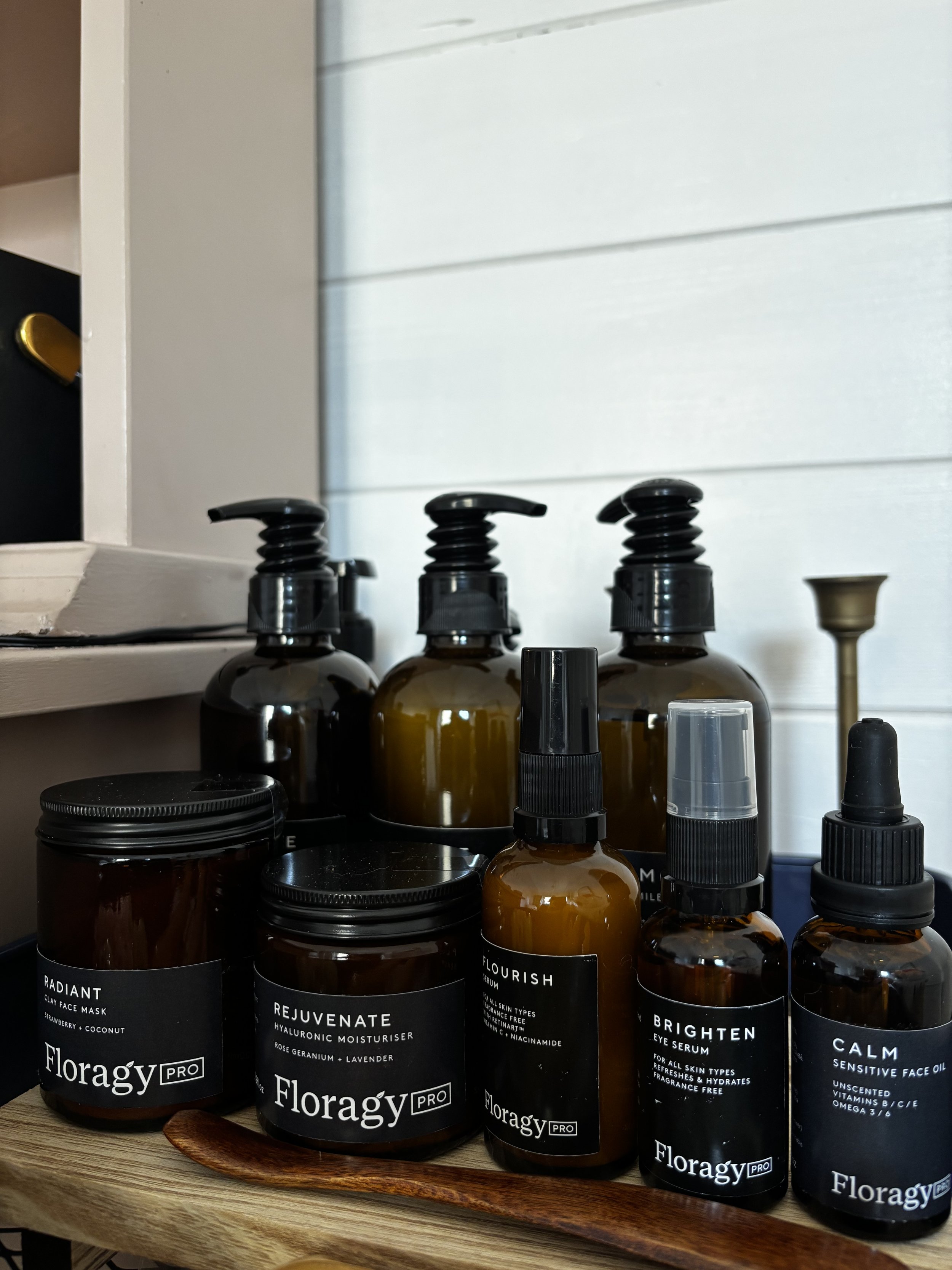 A collection of Floragy skincare products in amber bottles with black and clear pumps and droppers, labeled with names like Radiant, Rejuvenate, Flourish, Brighten, and Calm, placed on a wooden tray.