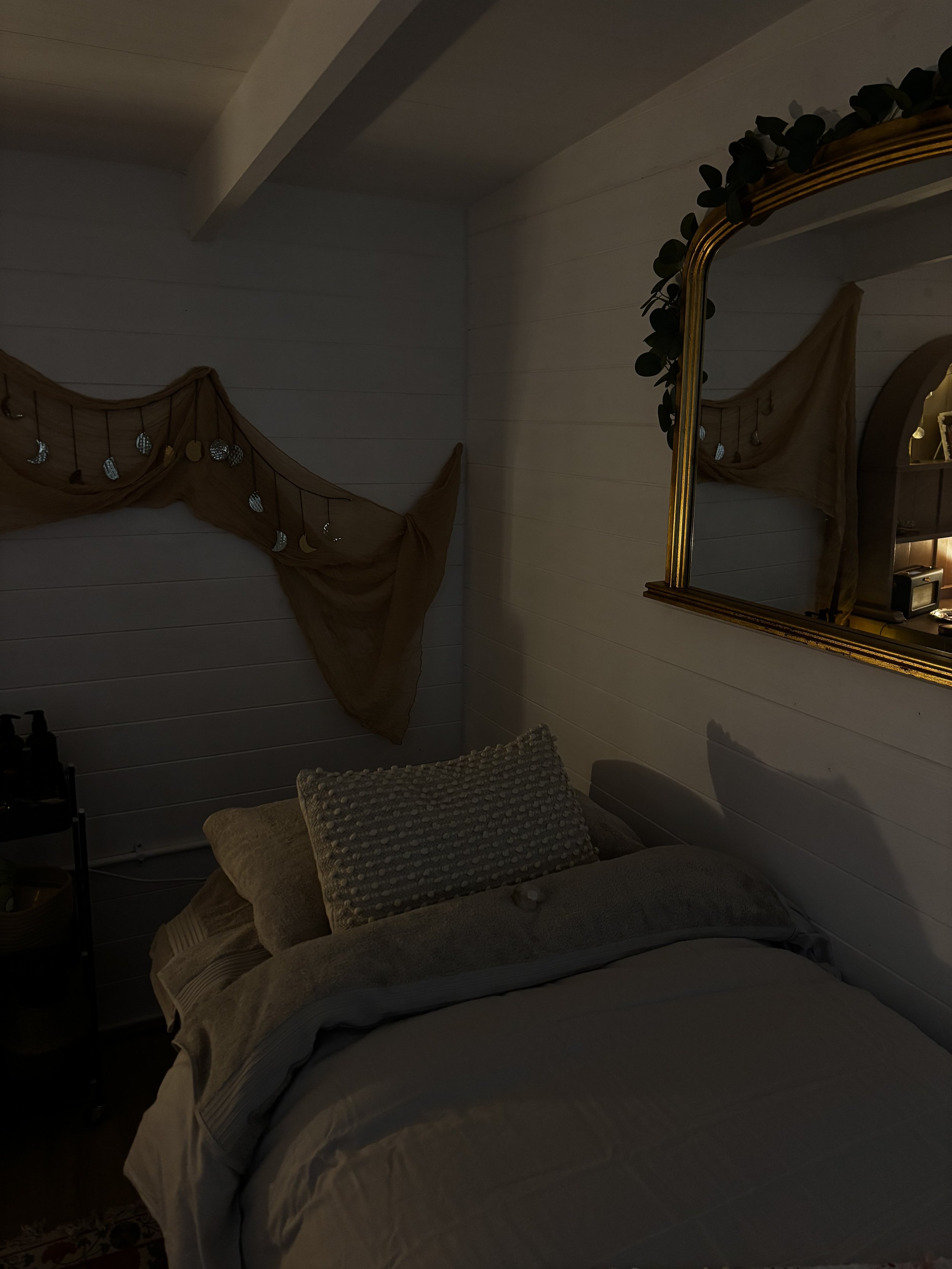 A cozy treatment room with a treatment couch, two pillows, a gold-framed mirror on the wall, and a decorative beige fabric with moon and star ornaments hanging on the wall, illuminated by soft lighting.
