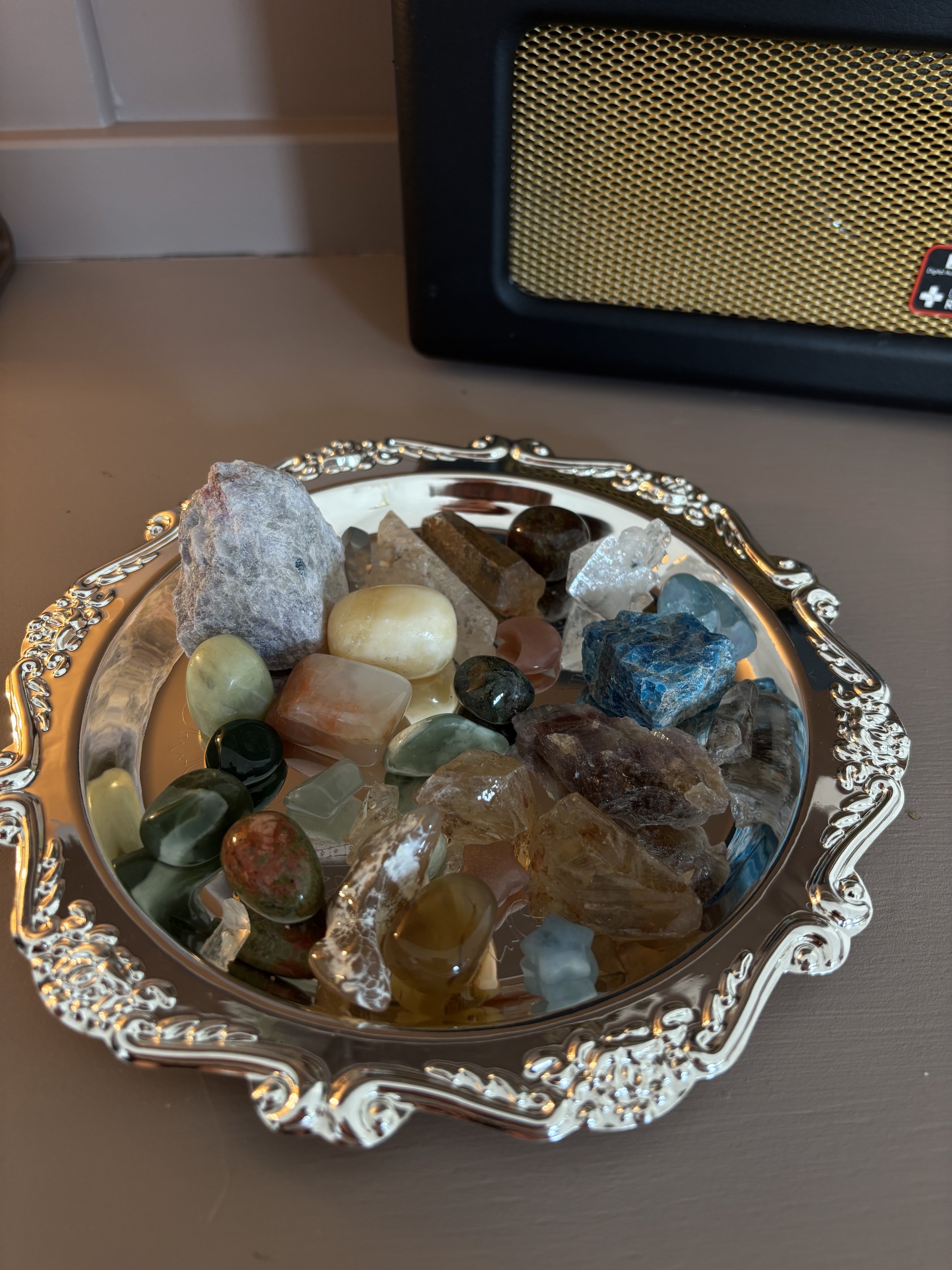 A decorative silver tray filled with various colorful polished and raw crystals, placed on a table with a black Roberts speaker in the background.