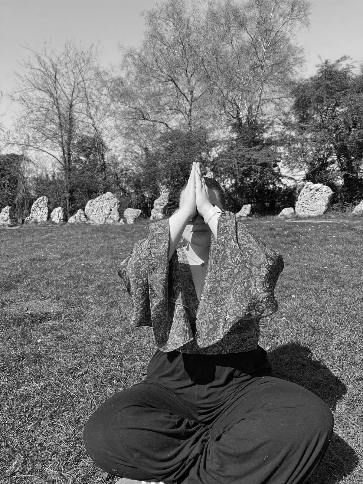 A woman sitting cross-legged on the grass outdoors, wearing a patterned jacket, with her hands pressed together in a prayer position covering her face, surrounded by trees and rocks in the background. Based at the rollright stones