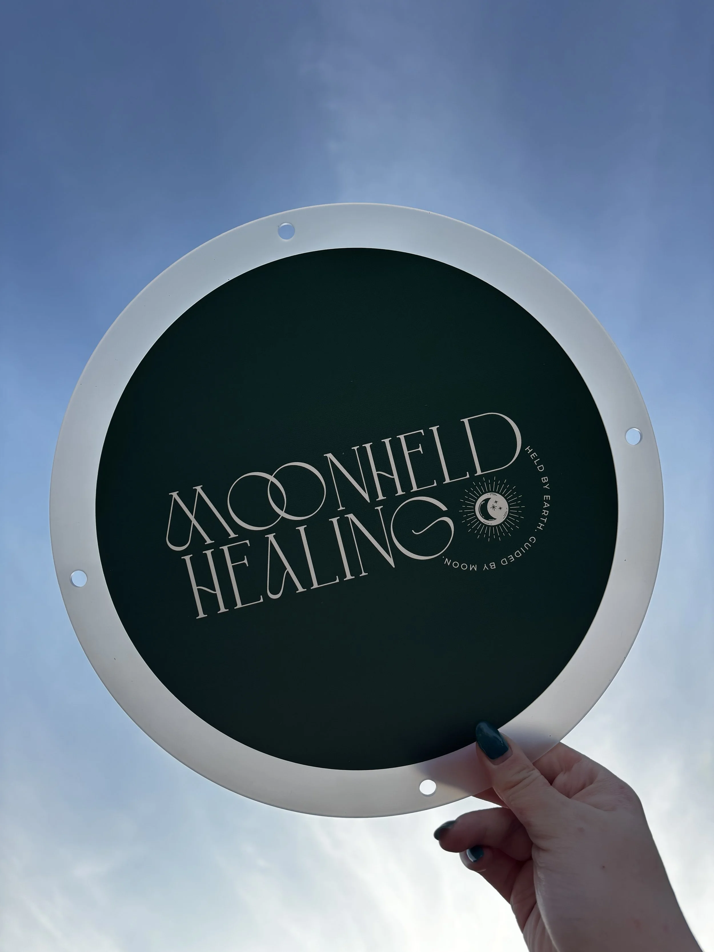 Person holding a circular sign that reads "Moonheld Healing" with a moon and star design, against a cloudy sky.
