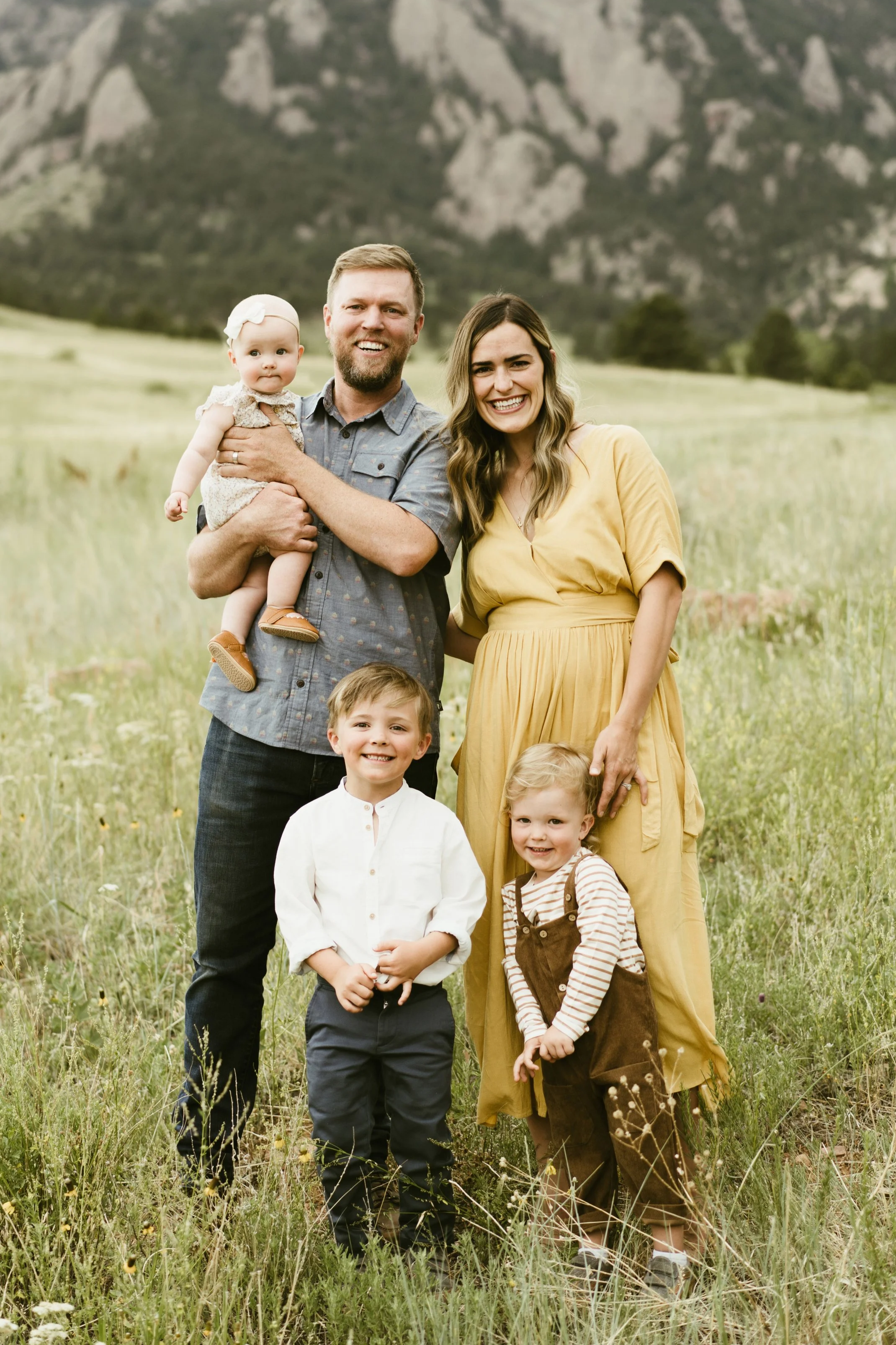 Family of five outdoors in a green field with mountains in the background, smiling at the camera.