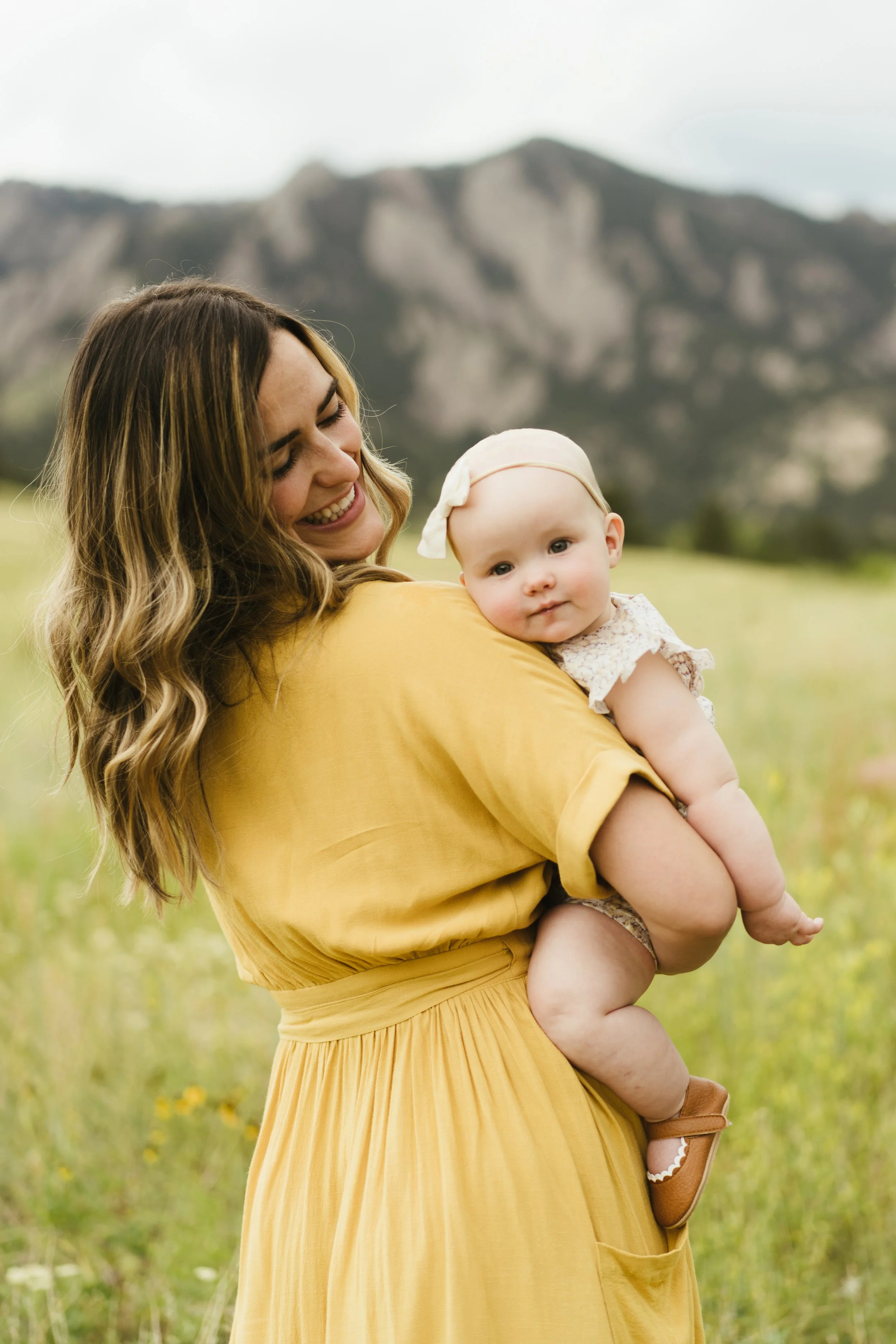 A woman with long wavy brown hair in a yellow dress holding a baby girl in a white dress with a bow headband in a green field with mountains in the background.