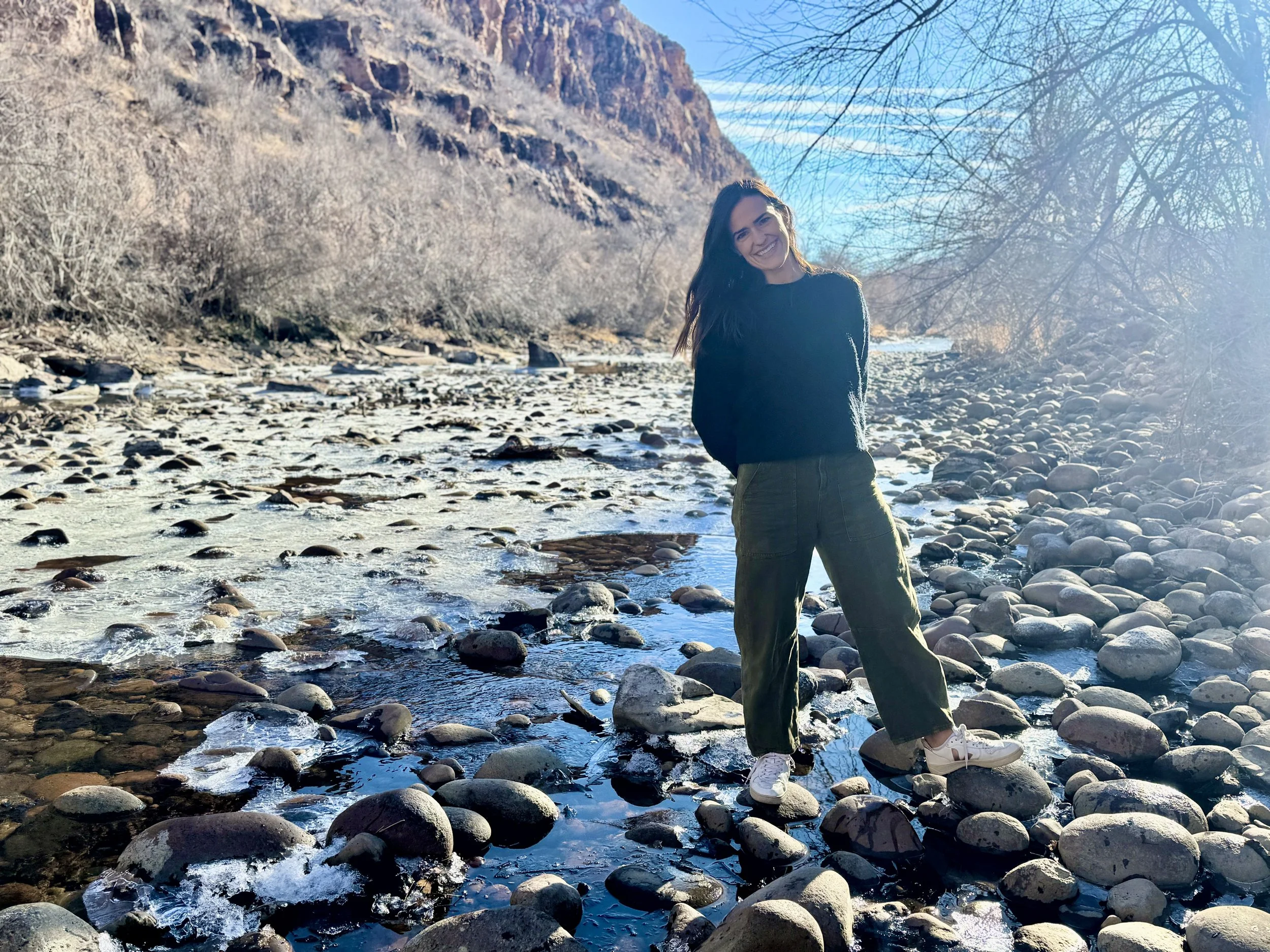 A smiling woman standing on a rocky riverbank with trees and mountains in the background on a clear, sunny day.