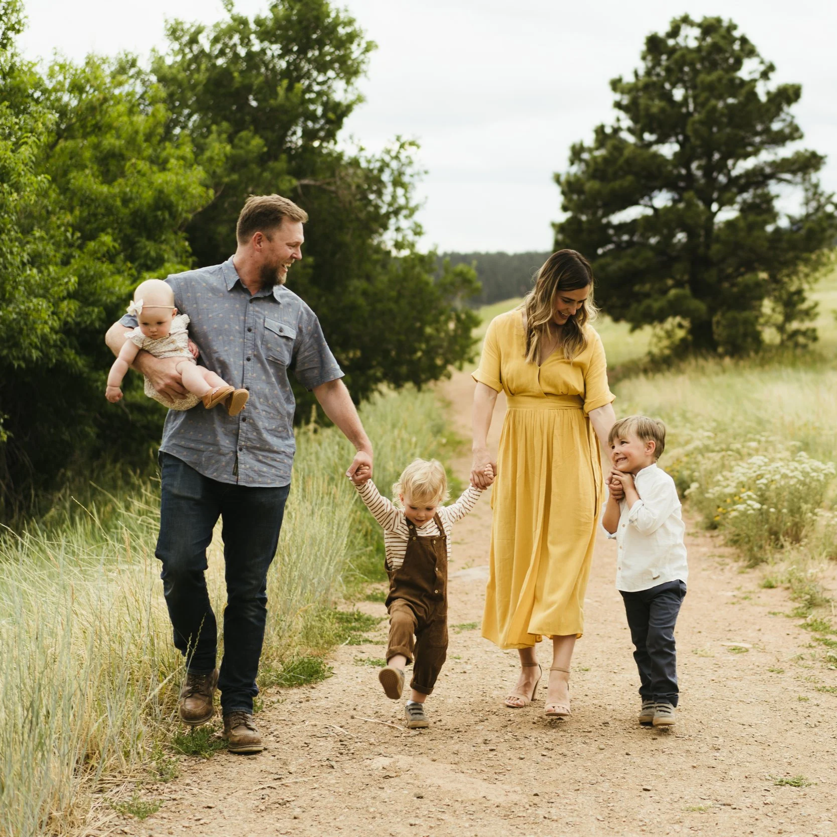 A family of five walking on a dirt path in a grassy and wooded area, holding hands and smiling.