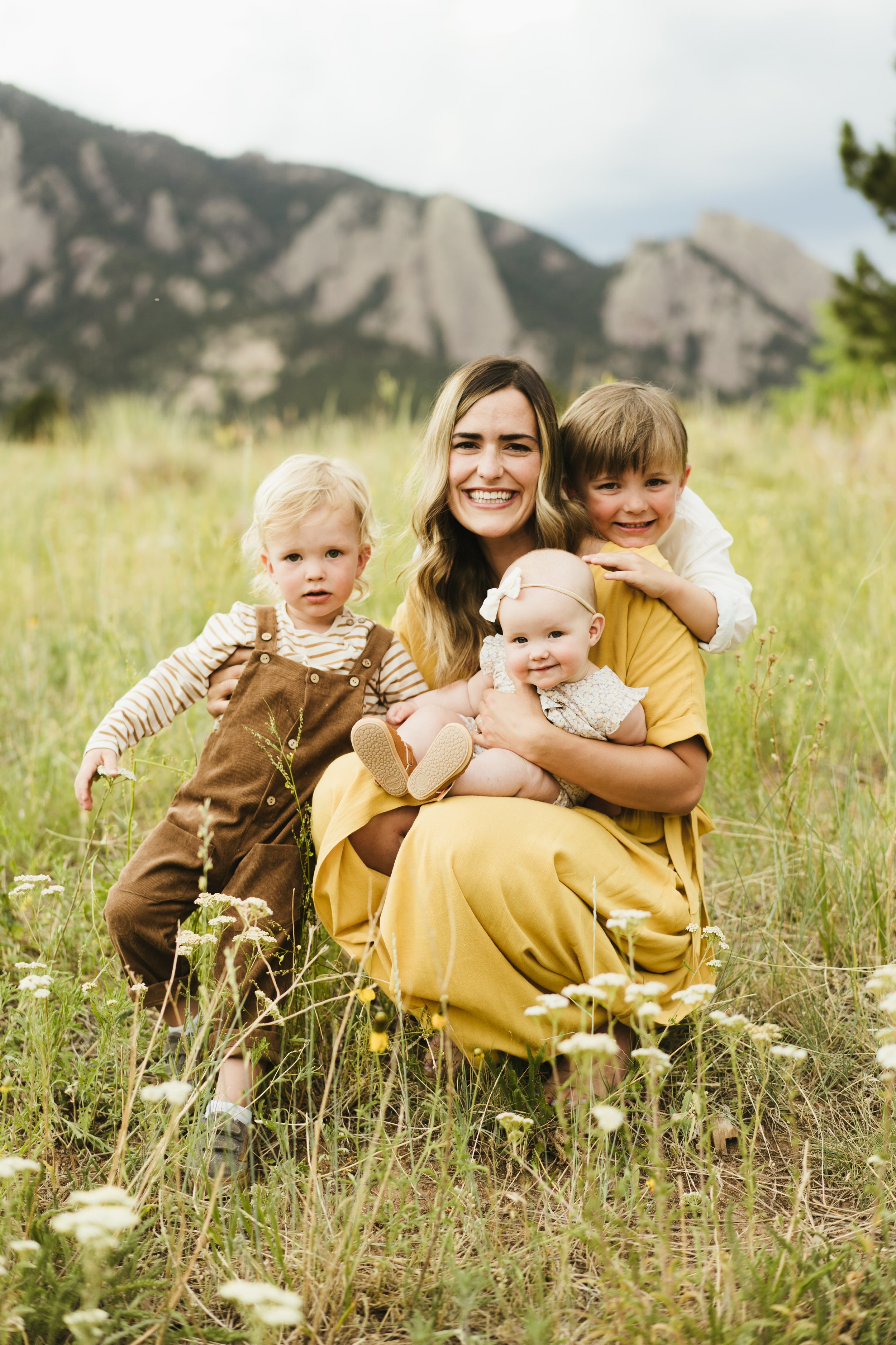 A woman with three young children in a grassy field with mountains in the background. The woman is smiling, holding a baby girl, while an older boy hugs her and a young girl stands nearby.