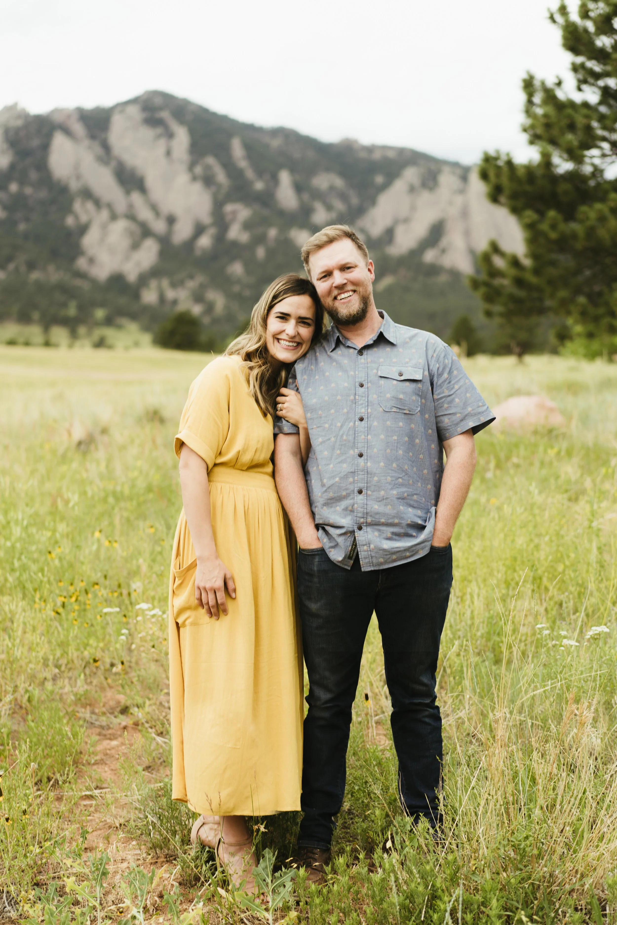 A smiling couple standing in a grassy field with mountains in the background, the woman in a yellow dress and the man in a short-sleeved button-up shirt.