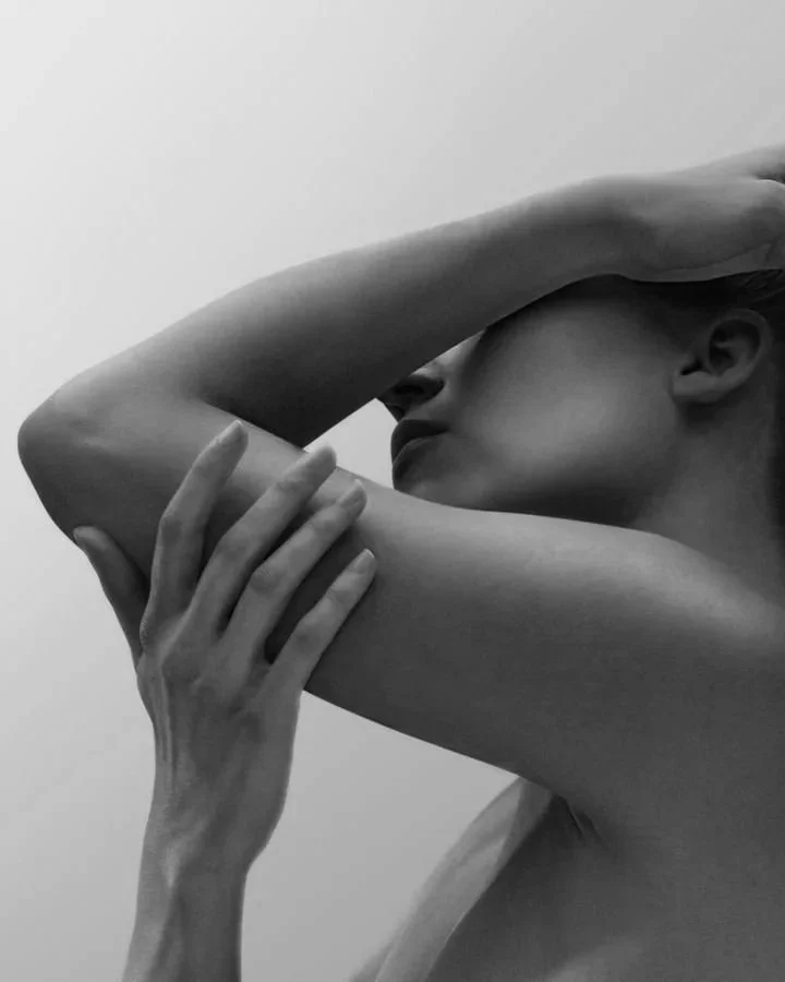 A black-and-white close-up photo of a woman with her arm bent, touching her forehead, with her other hand gently resting on her arm, emphasizing her smooth skin and facial profile.