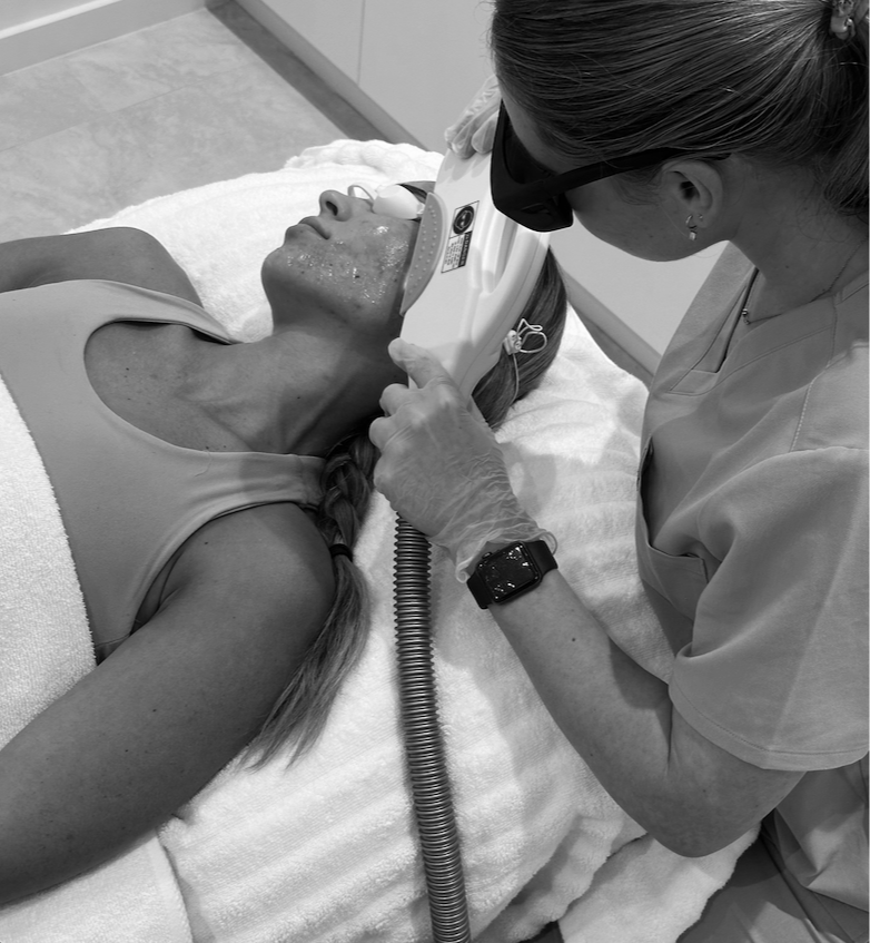 A medical professional in scrubs and gloves is performing a skincare treatment on a woman lying on a bed. The patient has a facial mask or treatment applied to her face, and the professional is using a device on her chin area.