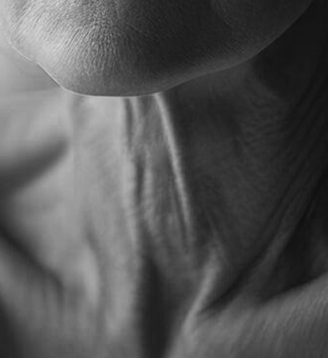 Close-up black and white photo of a person's neck and part of their chin, with a focus on textured skin and clothing.
