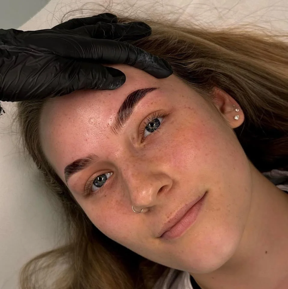 A young woman with light skin, blue eyes, and freckles receives an eyebrow grooming treatment from a person wearing black gloves.