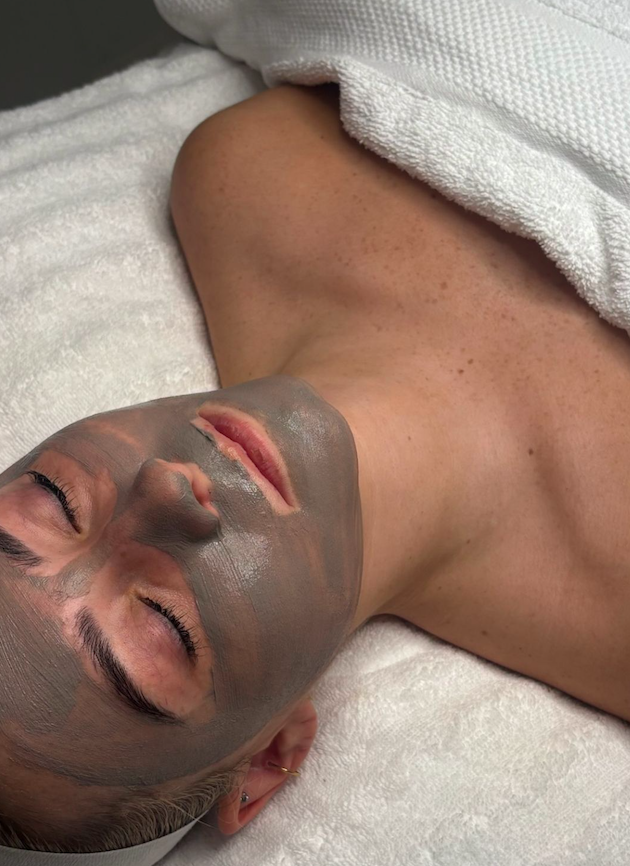 Person lying down with a gray facial mask, eyes closed, on a white towel or blanket.