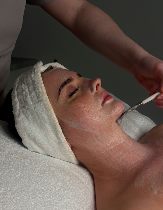 A woman receives a facial treatment while lying on a spa bed with her eyes closed, her head wrapped in a towel.