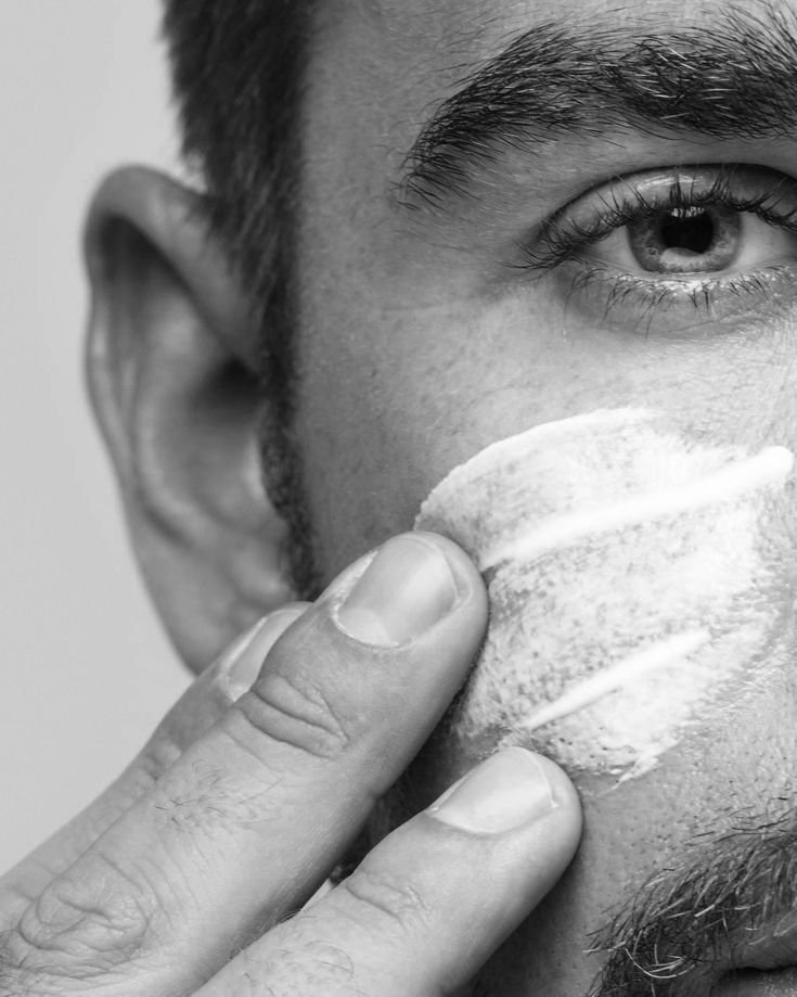 A close-up of a man with a trimmed beard applying shaving foam to his cheek with his fingers.