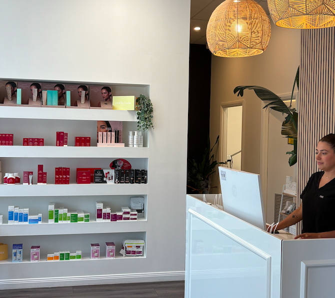 Interior of a modern skincare or beauty store with a white wall display shelf filled with skincare products organized by color, and a reception desk with a smiling staff member standing behind it. Hanging pendant lights and a large plant add to the decor.
