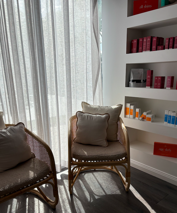 Sunlit corner in a beauty or skincare clinic with two rattan chairs, each with beige cushions, near white curtains and a shelf holding skincare products.