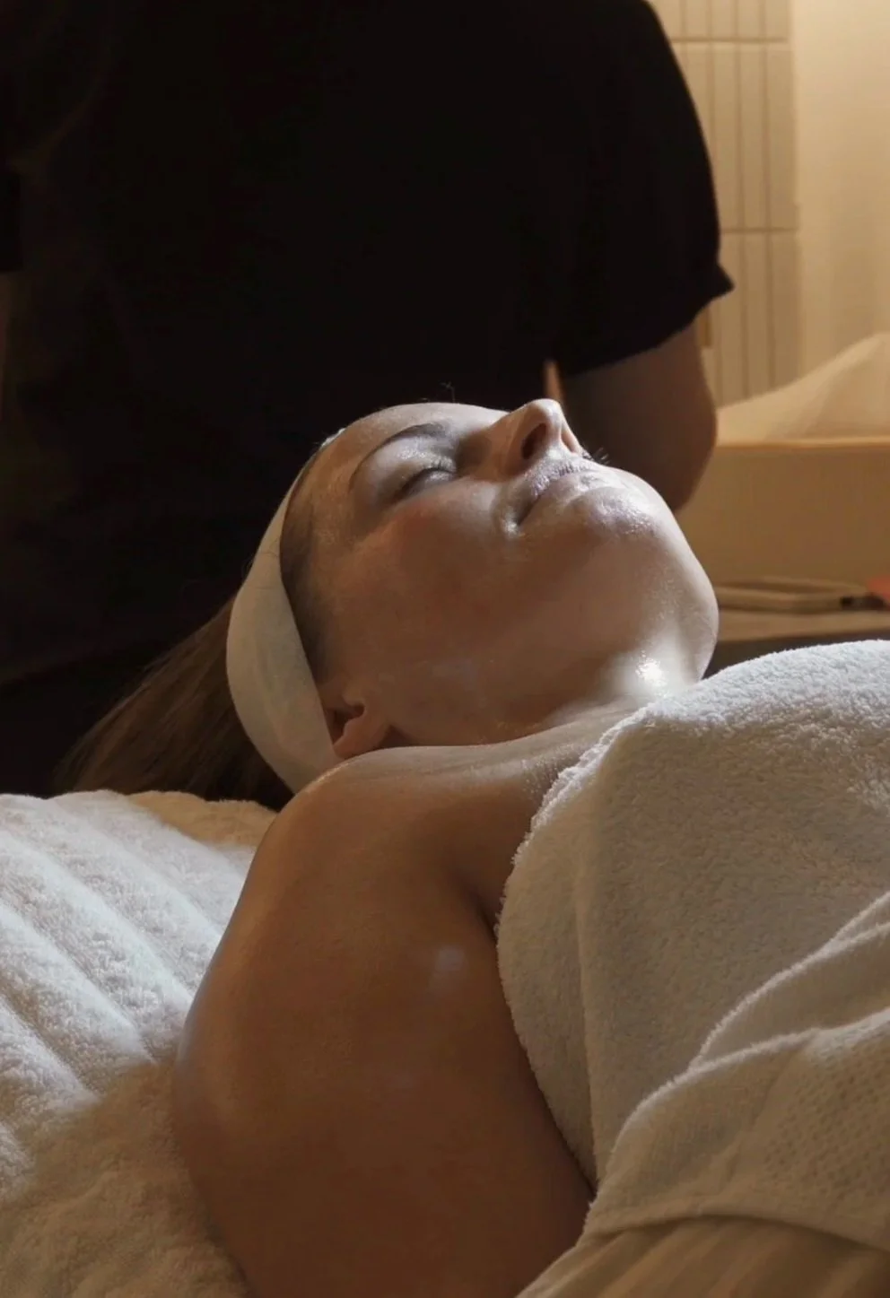 A woman receiving a facial treatment at a spa, lying peacefully with her eyes closed, wearing a white headband and towel, in a relaxing setting.