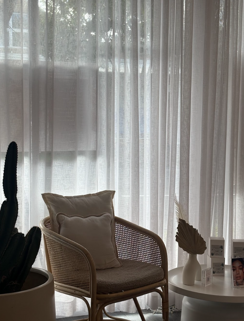 A cozy interior corner with a rattan chair holding a cream-colored cushion, beside a white round table with decorative objects, and sheer curtains allowing natural light to fill the space.