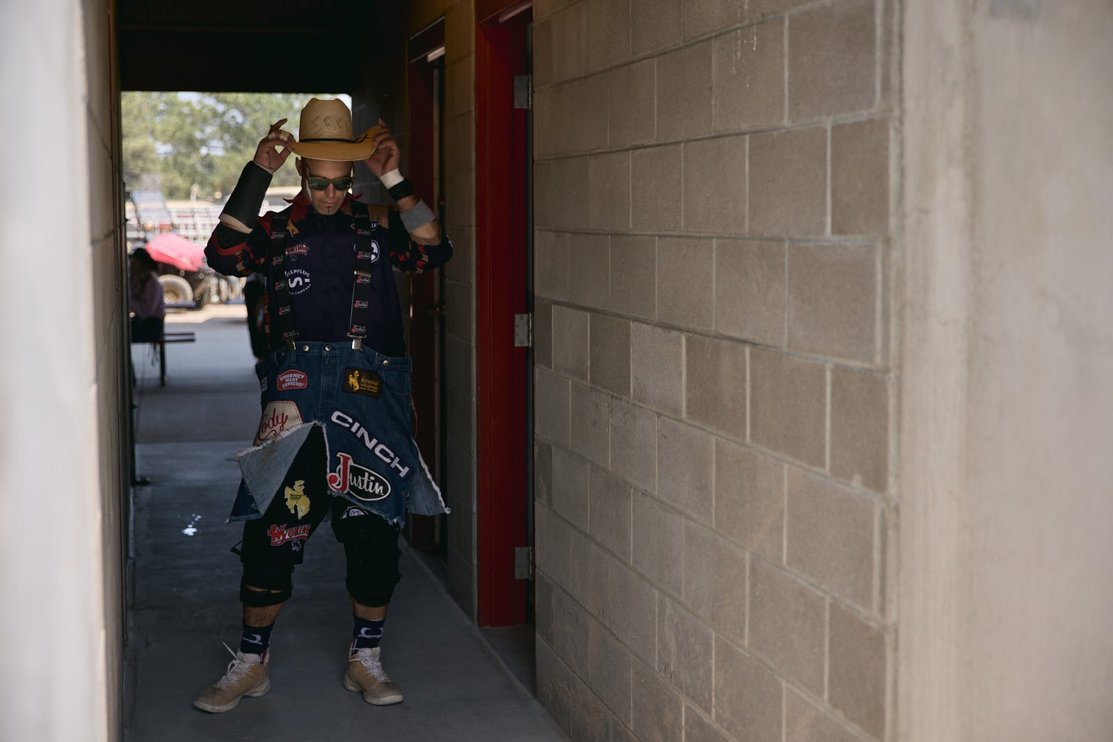 Rodeo clown adjusting a straw hat in a corridor, wearing a rodeo clown suit with sponsor logos and patches.