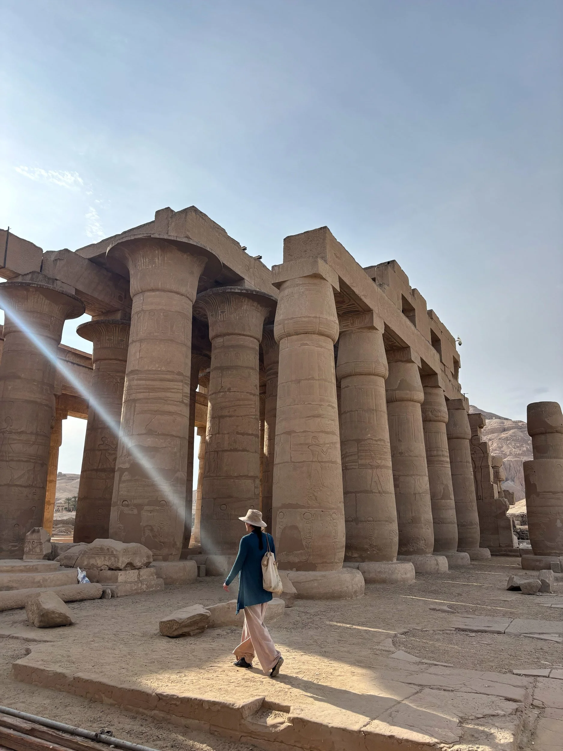 A woman in a blue long sleeve top, pink pants, and a wide-brimmed hat walking among ancient Egyptian stone columns at an archaeological site. The columns have hieroglyphic carvings, and the sky is clear with sunlight streaming into the scene.