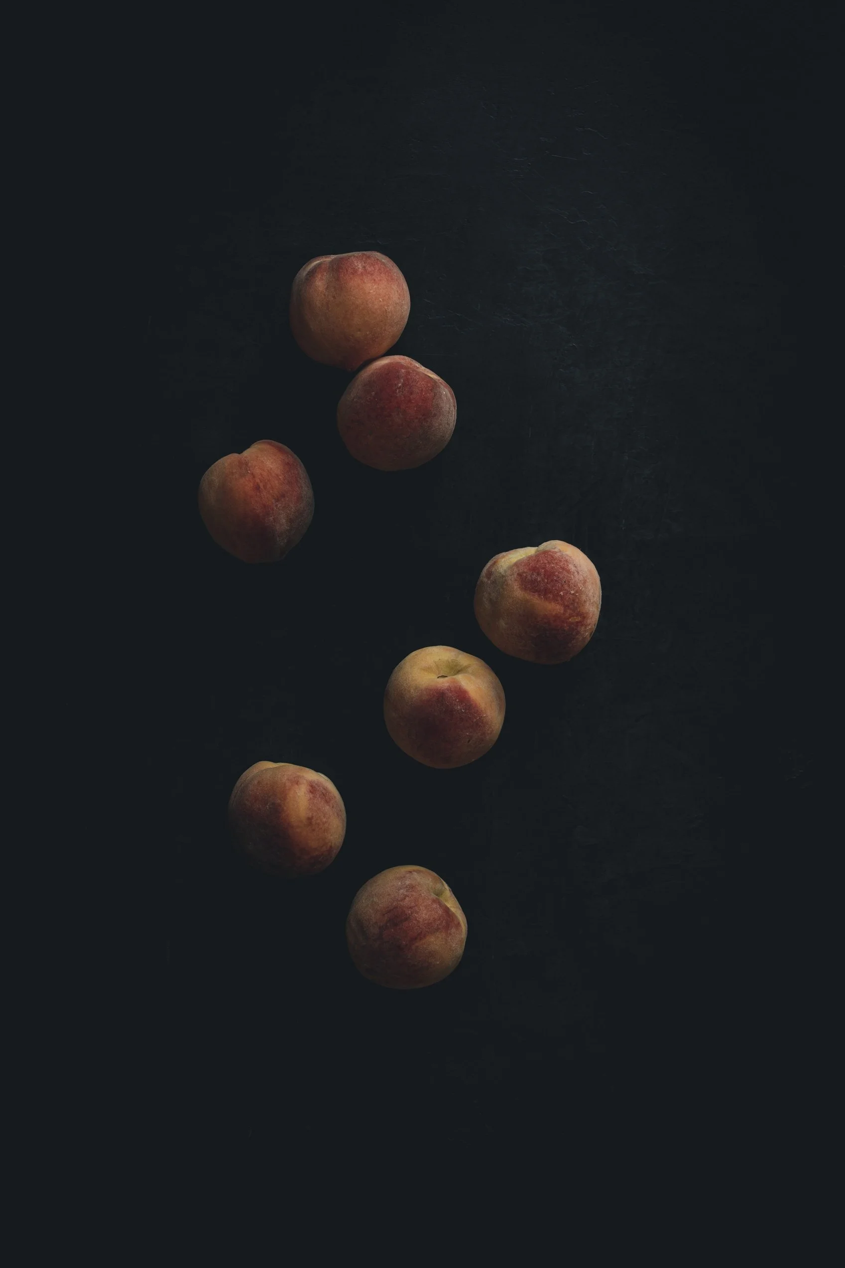 Eight peaches scattered on a dark surface, with soft, directional lighting.