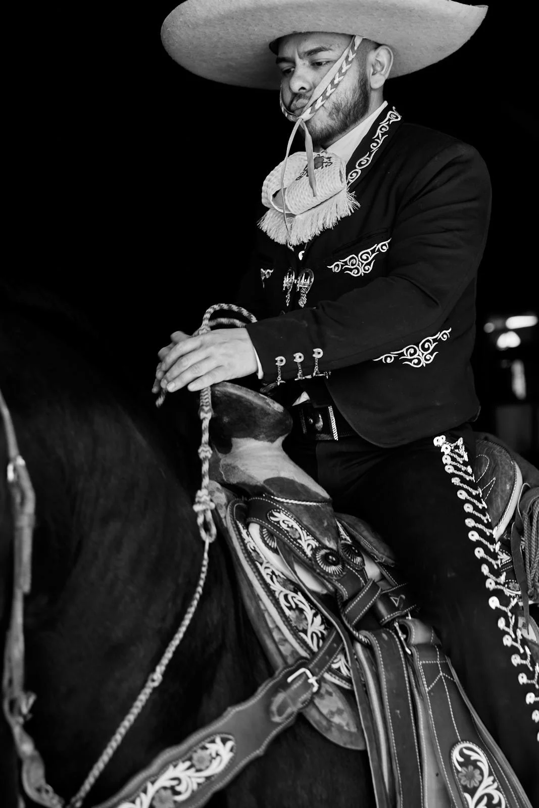 A man dressed in traditional Mexican attire, wearing a large sombrero and traditional embroidered suit, riding a horse, with a decorative saddle in black and white.