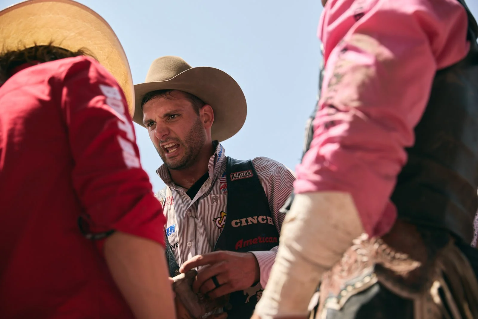 A man with a cowboy hat and striped shirt appears to excited, surrounded by two people wearing pink and red shirts, outdoors under a clear blue sky.