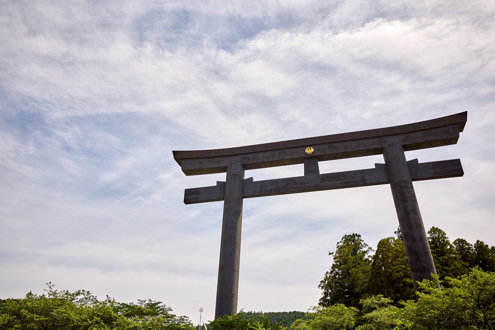 A traditional wooden Japanese torii gate set against a partly cloudy sky with green trees in the foreground.