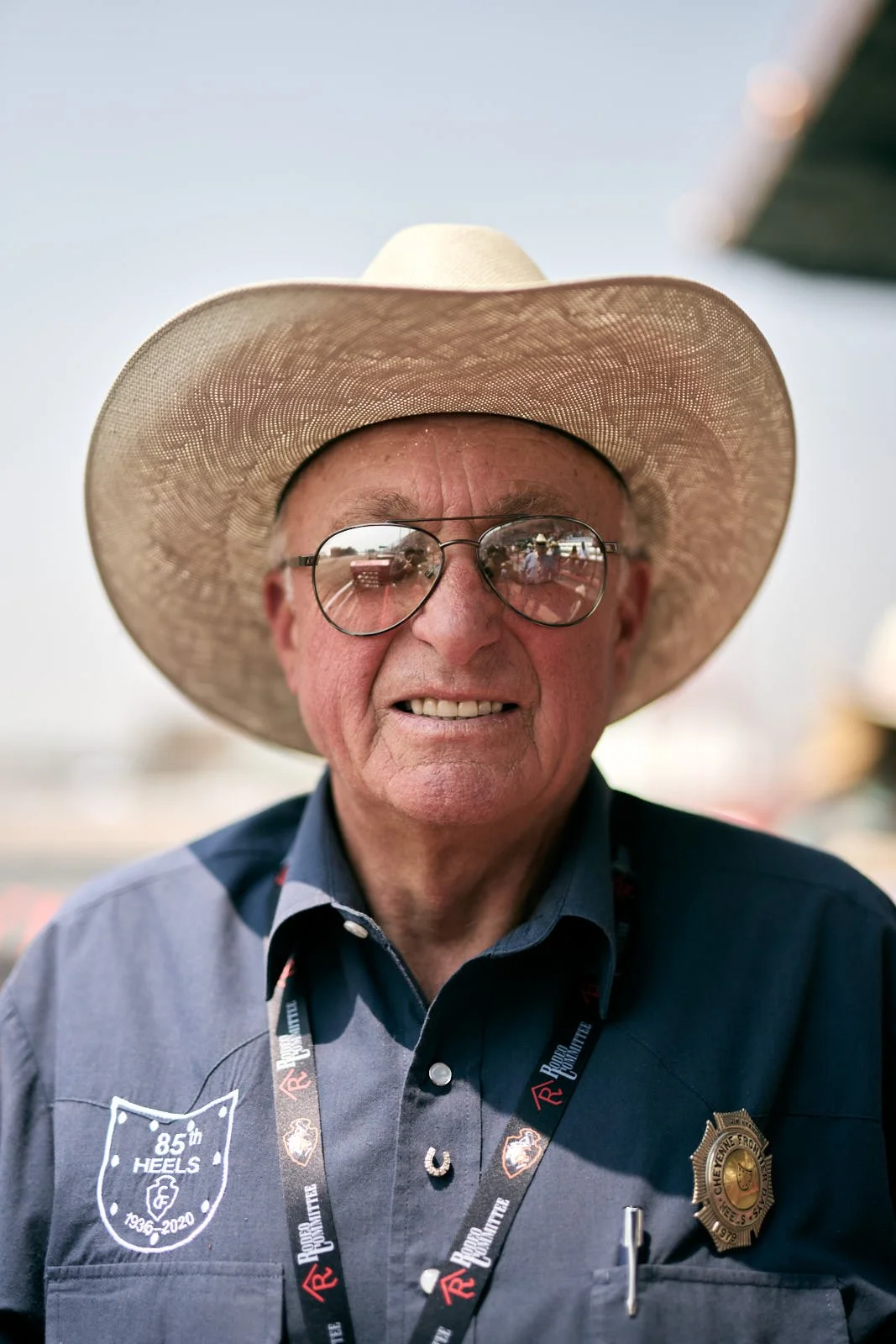An elderly man wearing a large straw cowboy hat, sunglasses, a dark blue shirt, and a badge, smiling at the camera.
