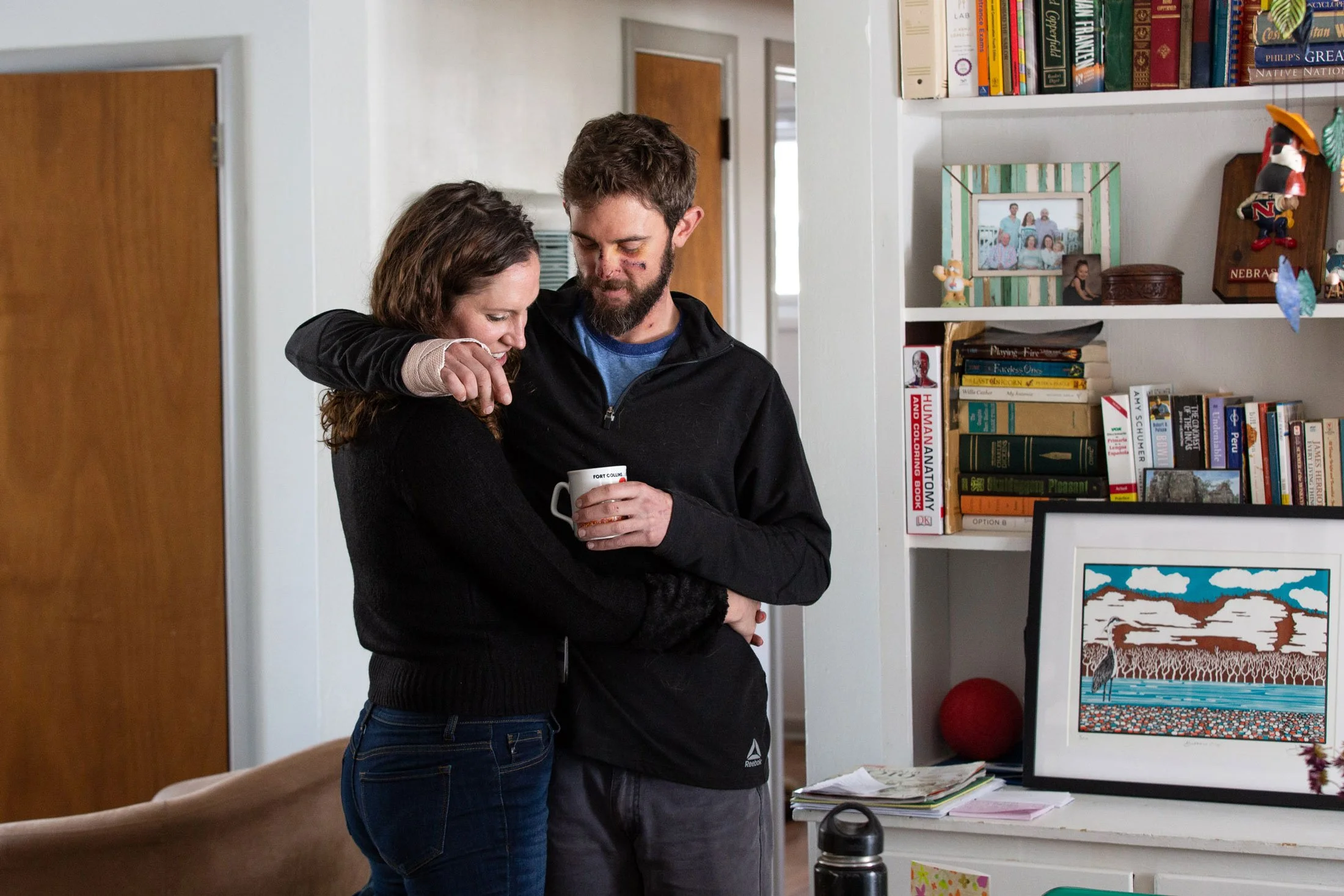 A man and woman hugging inside a home, smiling. The man is holding a coffee mug, and the woman is resting her head on his shoulder. They are standing near a bookshelf filled with books and decorations.