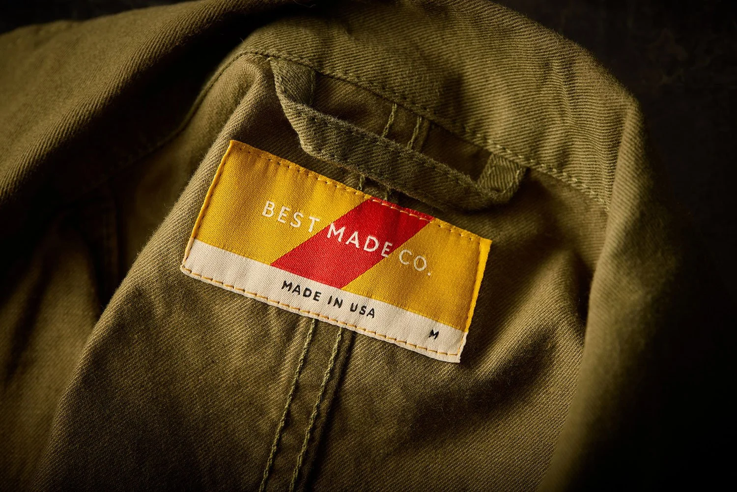 Close-up of a clothing label on an olive green jacket, with the brand name 'BEST MADE CO.', 'MADE IN USA', and a red, yellow, and white striped design.