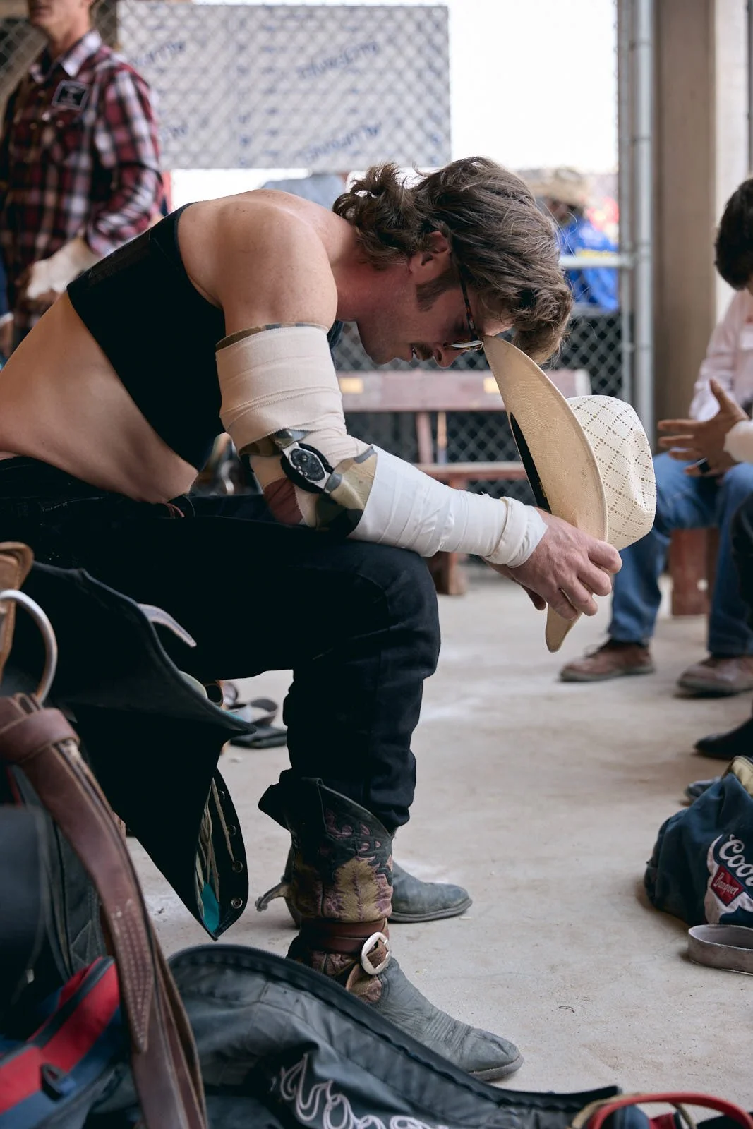 A man with long hair and glasses, sitting on a bench, wearing a black tank top, with a padded arm, holding a cowboy hat.