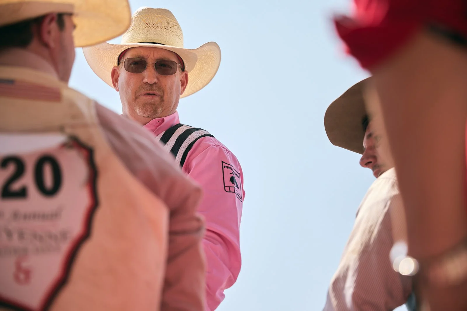 A man with sunglasses and a wide-brimmed hat speaking to a group, seen from below with a clear blue sky in the background.