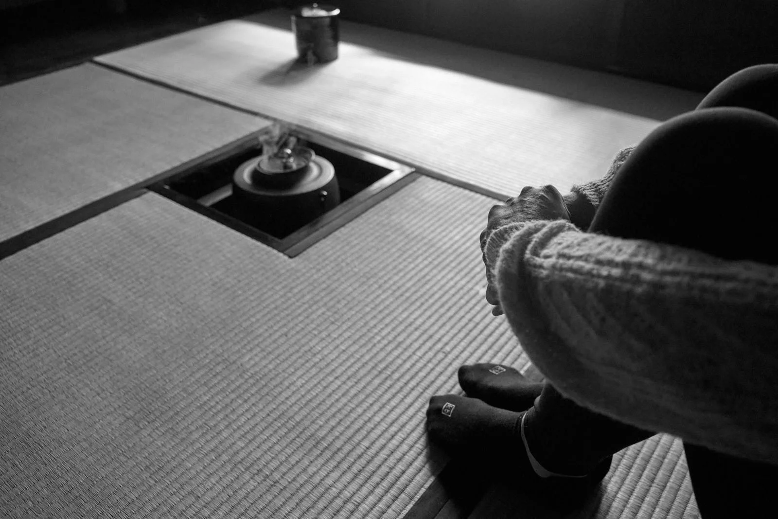 A person sitting on a tatami mat with their hands clasped and head bowed, in a traditional Japanese setting. A small tea kettle is placed on the floor.